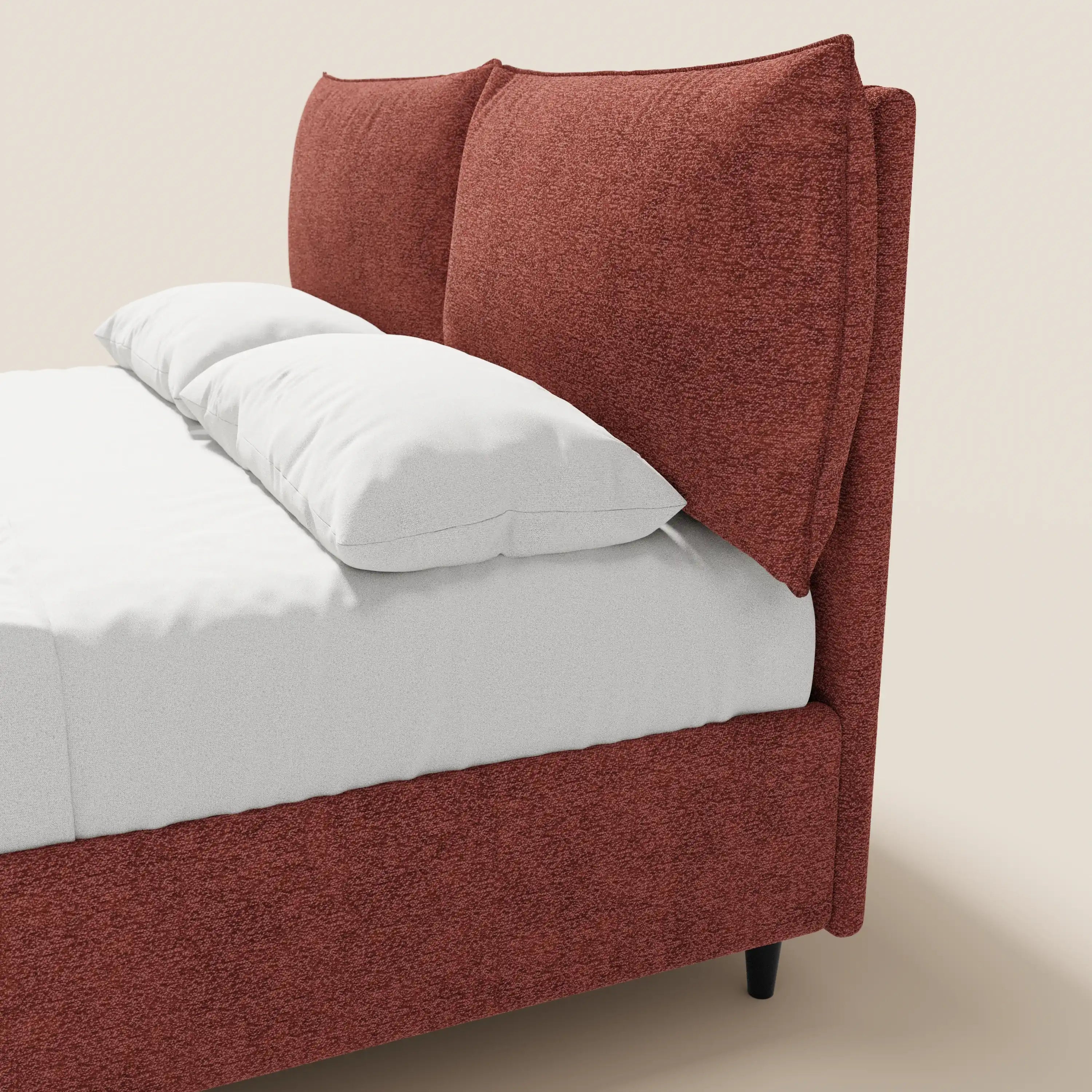 Un primo piano del letto imbottito ZAYRON di Letti.store, caratterizzato da una struttura in tessuto color ruggine certificato Oeko-Tex® e da ampi cuscini regolabili. Il letto è rifatto con cura con biancheria da letto bianca, su uno sfondo beige chiaro a tinta unita.