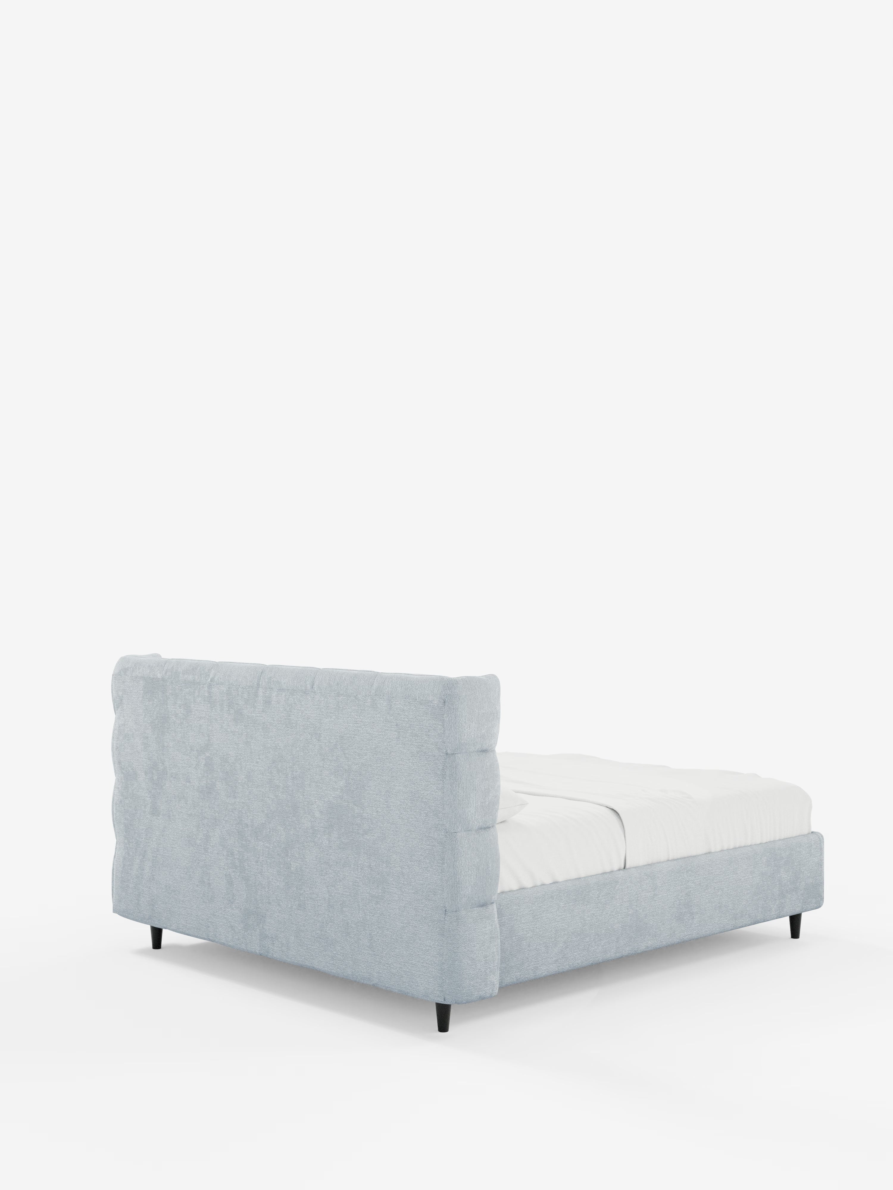 Un letto imbottito RAGNAR grigio chiaro con contenitore e testiera capitonné avvolgente di Letti.store, realizzato in elegante tessuto certificato Oeko-Tex®, è esposto su uno sfondo bianco. Il letto presenta una testiera alta e trapuntata e gambe corte di colore nero; è rivestito con un lenzuolo con angoli bianco ed è ripreso da un’angolazione posteriore sinistra.