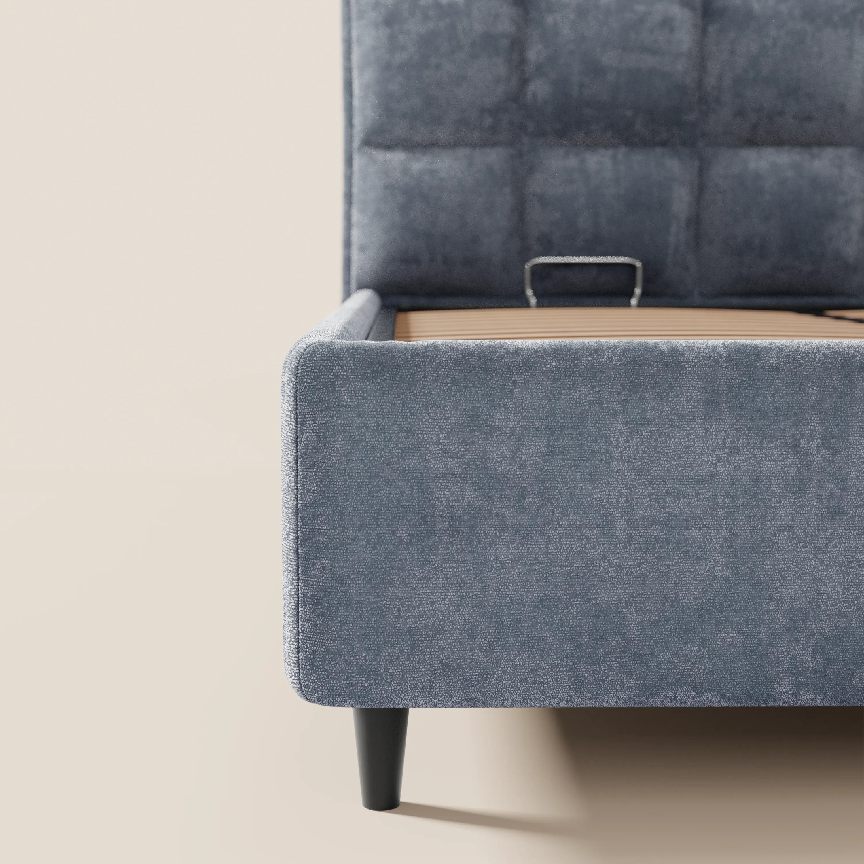 Primo piano del letto imbottito AURO di Letti.store, caratterizzato da un'alta testiera capitonné in ciniglia blu-grigia certificata Oeko-Tex® e da gambe affusolate nere. La struttura moderna del letto include un vano contenitore ed è raffigurata su uno sfondo bianco sporco.