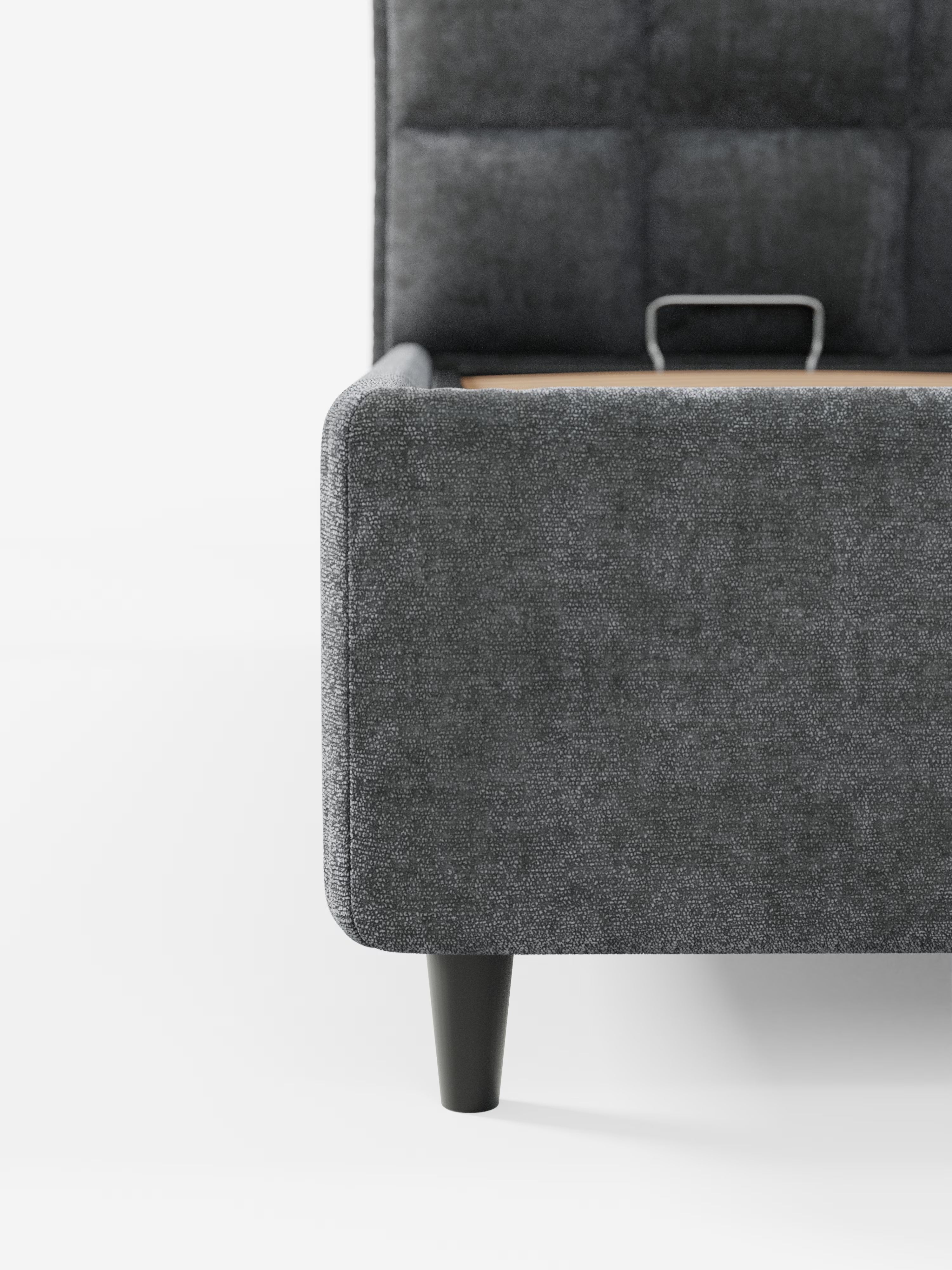 Primo piano del letto imbottito AURO con vano contenitore e testiera capitonné alta in ciniglia Oeko-Tex® di Letti.store, qui raffigurato con rivestimento in grigio moderno. Su uno sfondo bianco a tinta unita sono visibili solo l’angolo anteriore inferiore e una gamba nera affusolata, con una vista parziale della struttura metallica del vano contenitore all’interno del letto.