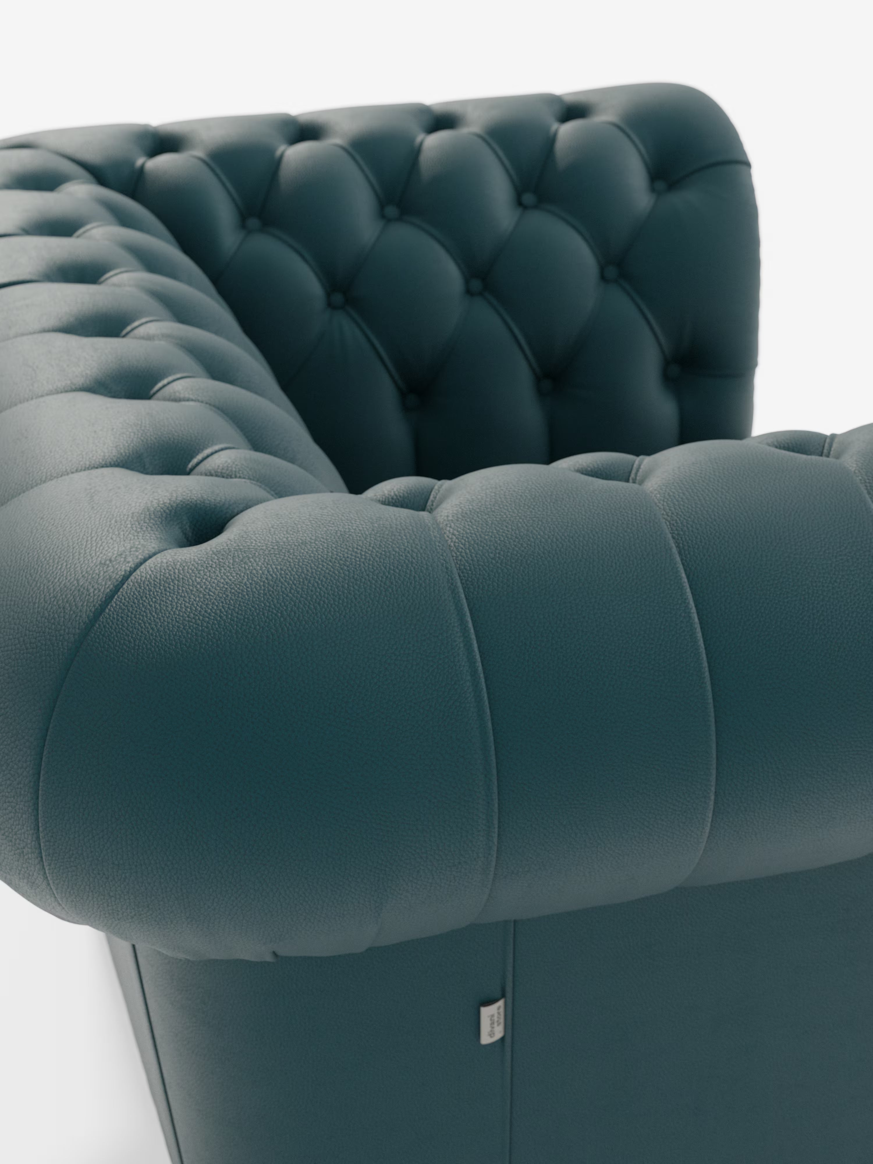 Primo piano dell'angolo della poltrona Chester in vera pelle spessorata con capitonné artigianale di Divani.Store: una poltrona in pelle color verde acqua scuro con capitonné, ispirata al classico design della poltrona capitonné, caratterizzata da profondi bottoni decorativi. Il rivestimento in vera pelle, morbida e soffice, è impreziosito da una piccola etichetta metallica applicata sulla parte inferiore della poltrona. Immagine su sfondo bianco.