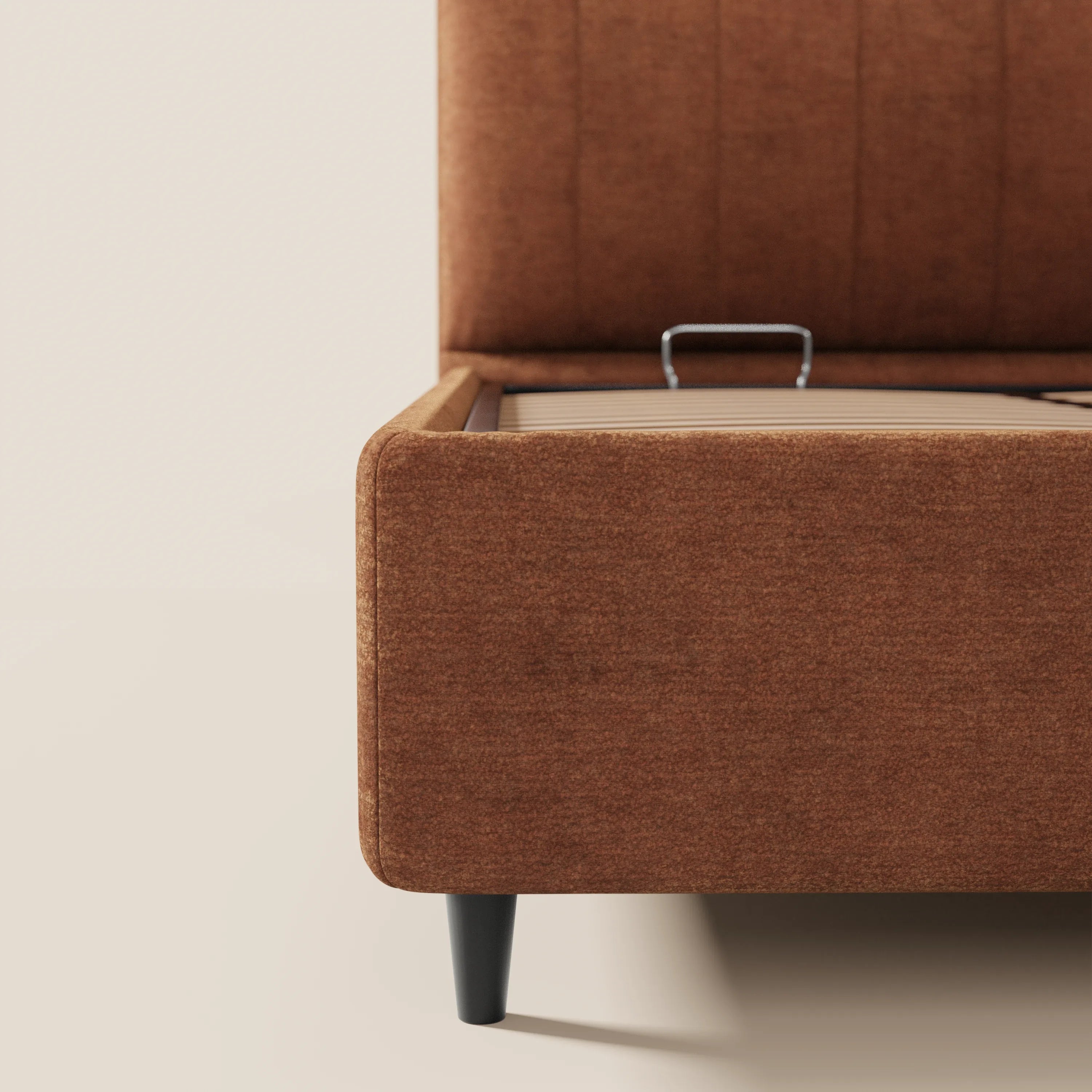 Primo piano del letto imbottito con contenitore ONYX di Letti.store, caratterizzato da gambe nere e da un vano contenitore aperto con maniglia in metallo. La parete e il pavimento color bianco sporco mettono in risalto il suo design moderno e minimalista. Tessuto certificato Oeko-Tex®.