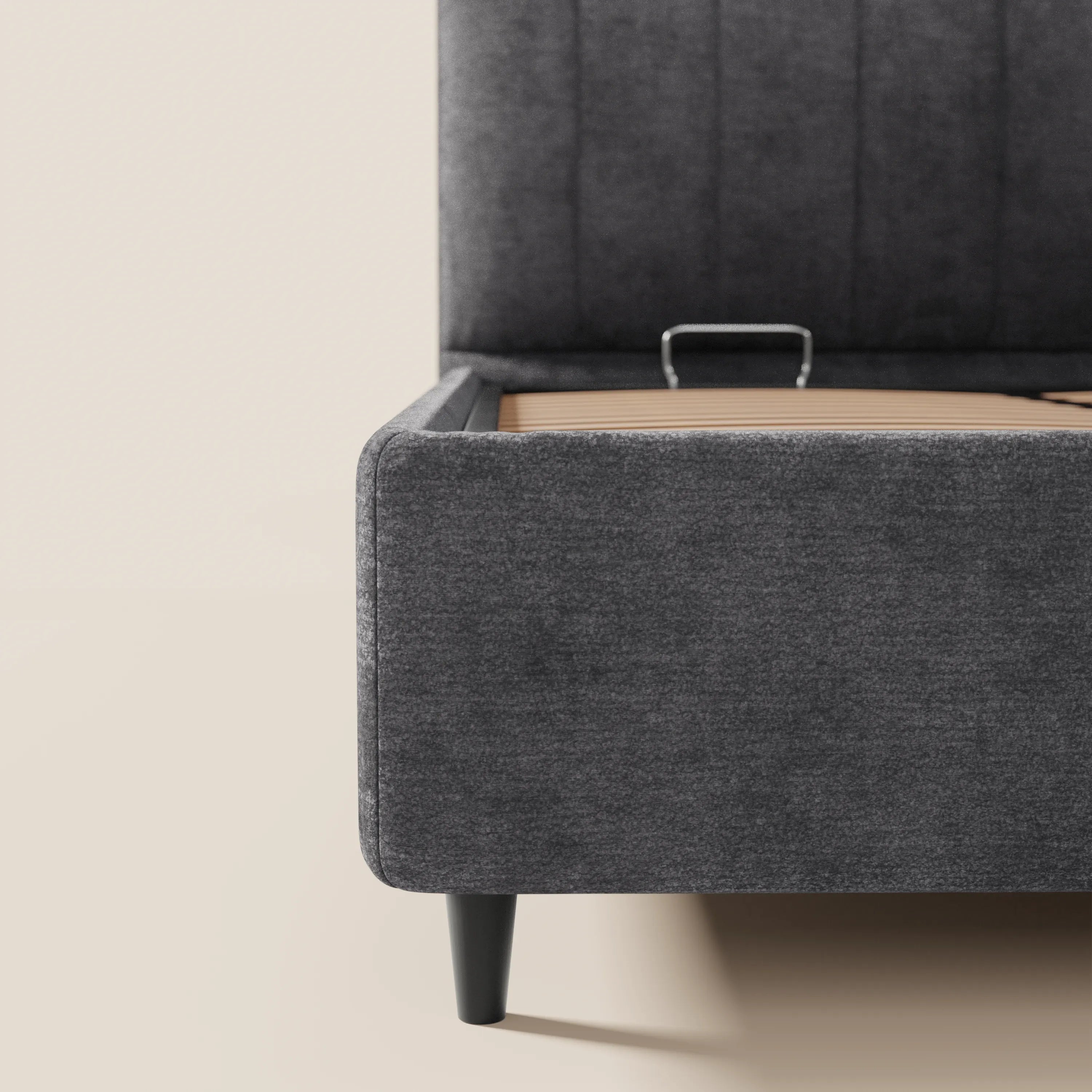 Un primo piano del letto imbottito ONYX di Letti.store, realizzato in tessuto grigio scuro certificato Oeko-Tex®, ne mette in risalto la morbida trama e il vano portaoggetti aperto con maniglia in metallo situato sotto il materasso, il tutto su uno sfondo beige chiaro.