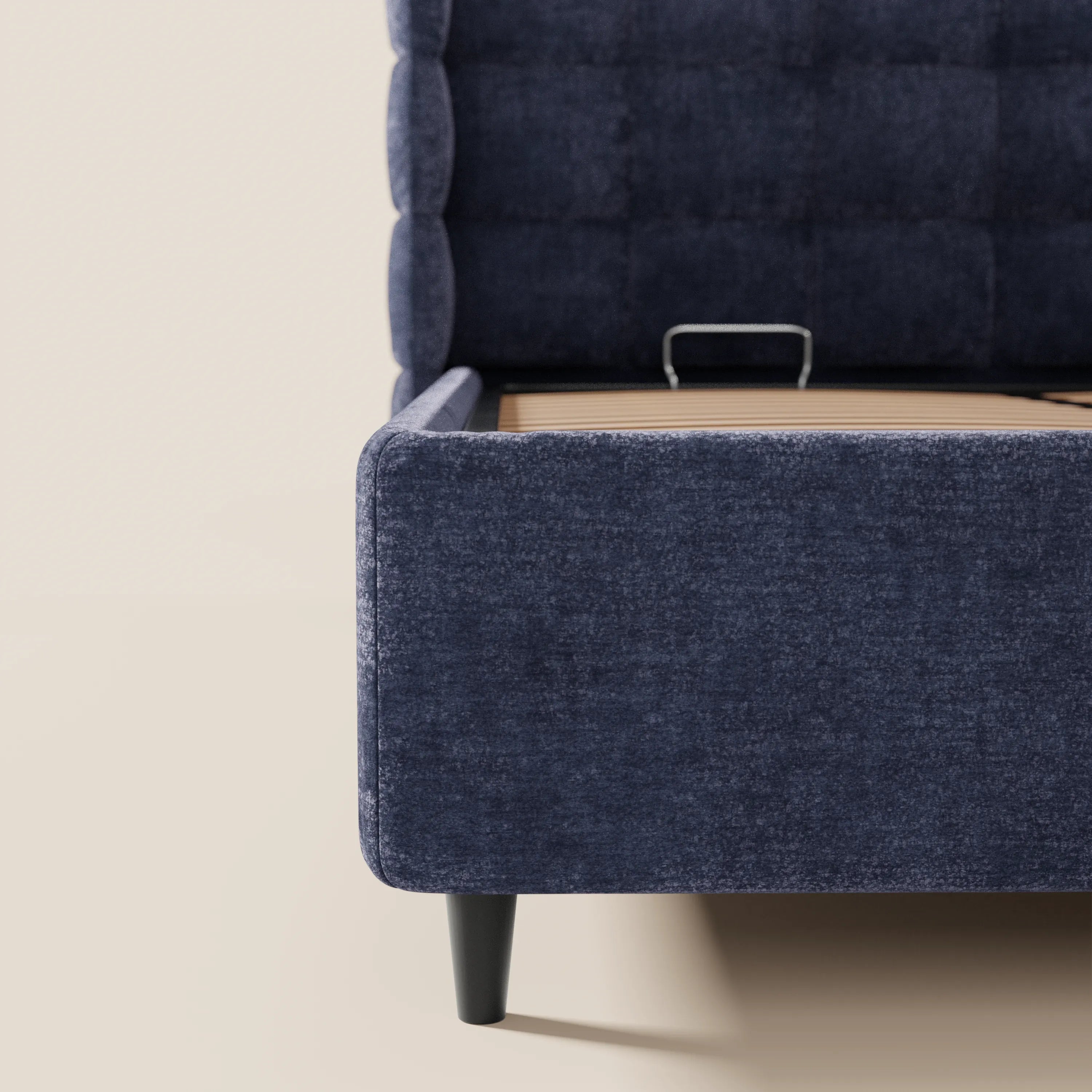 Primo piano dell'angolo del letto imbottito RAGNAR con contenitore e testiera capitonné avvolgente di Letti.store, realizzato in tessuto blu scuro certificato Oeko-Tex®, con gambe corte nere e una staffa di sostegno in metallo sulla base del letto, su uno sfondo beige chiaro.