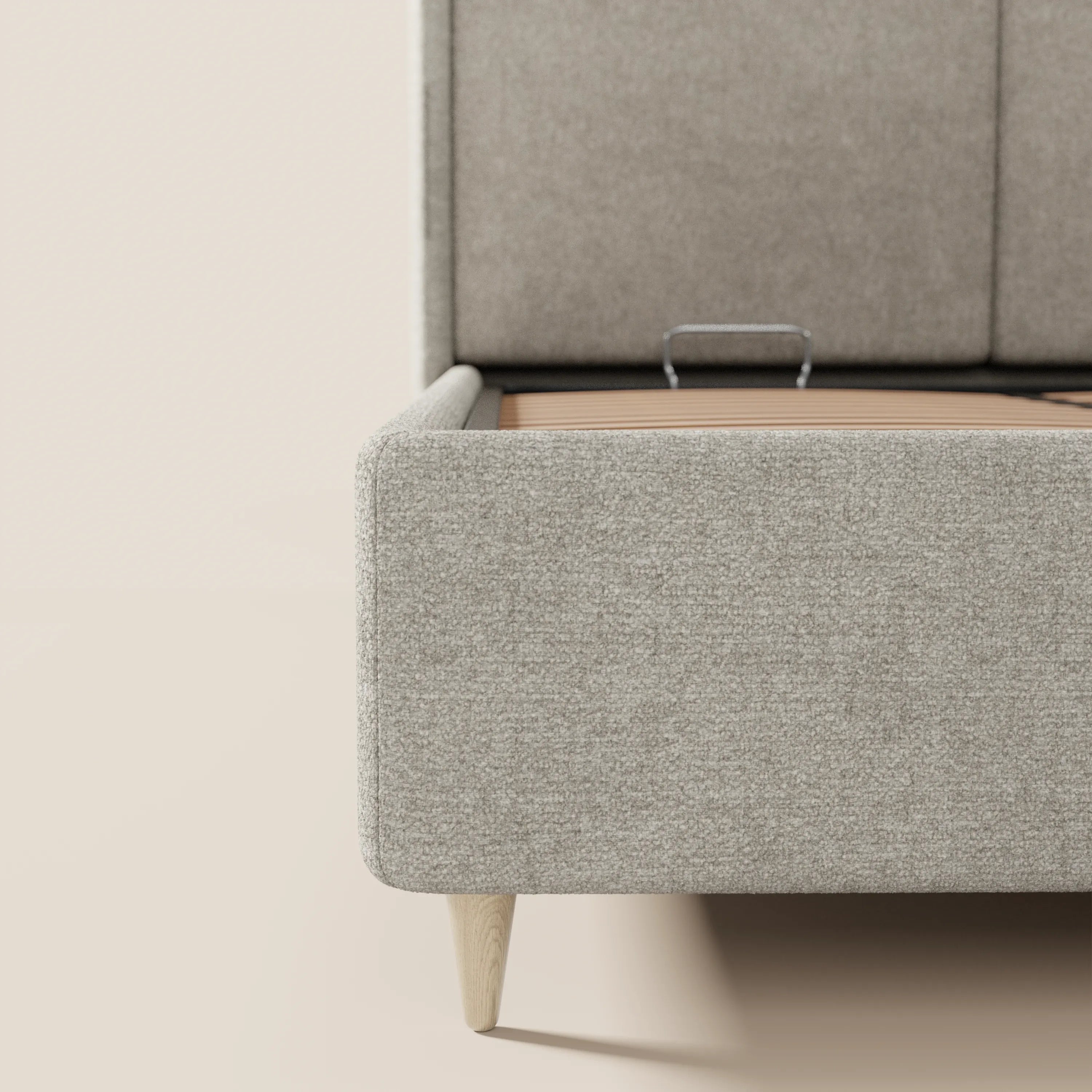 Primo piano del letto imbottito bicolore VALKIRIA di Letti.store, caratterizzato da un vano contenitore rivestito in beige con maniglia in metallo, da una caratteristica gamba in legno, da cuscini per lo schienale e da una testiera curva e avvolgente. Nell'immagine è visibile solo una parte del letto.