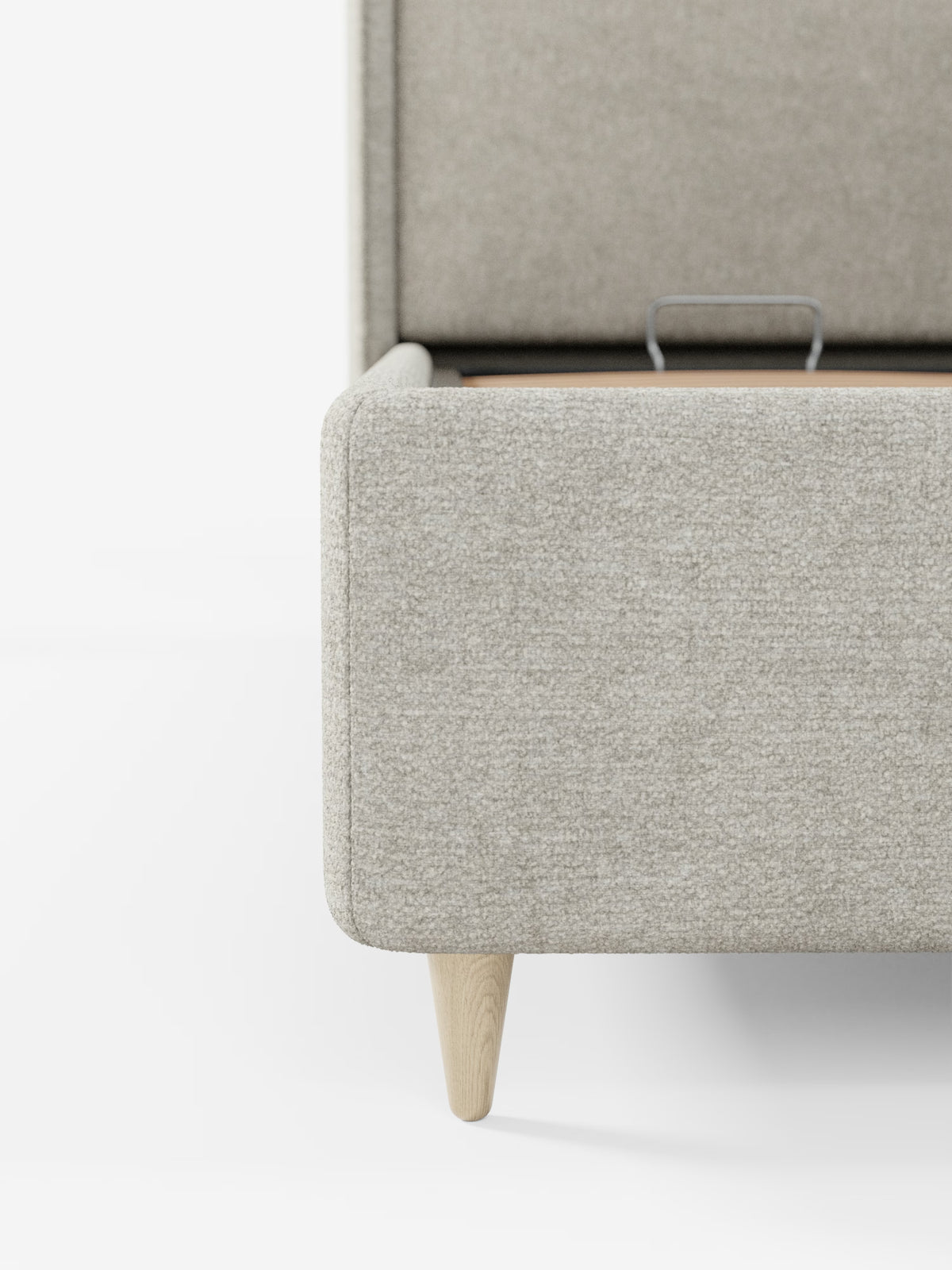 Primo piano dell'angolo del letto imbottito bicolore VALKIRIA con vano contenitore di Letti.store, caratterizzato da gambe in legno chiaro. La struttura imbottita presenta un pratico vano contenitore con il coperchio leggermente socchiuso, che lascia intravedere un'elegante maniglia in metallo su sfondo bianco.