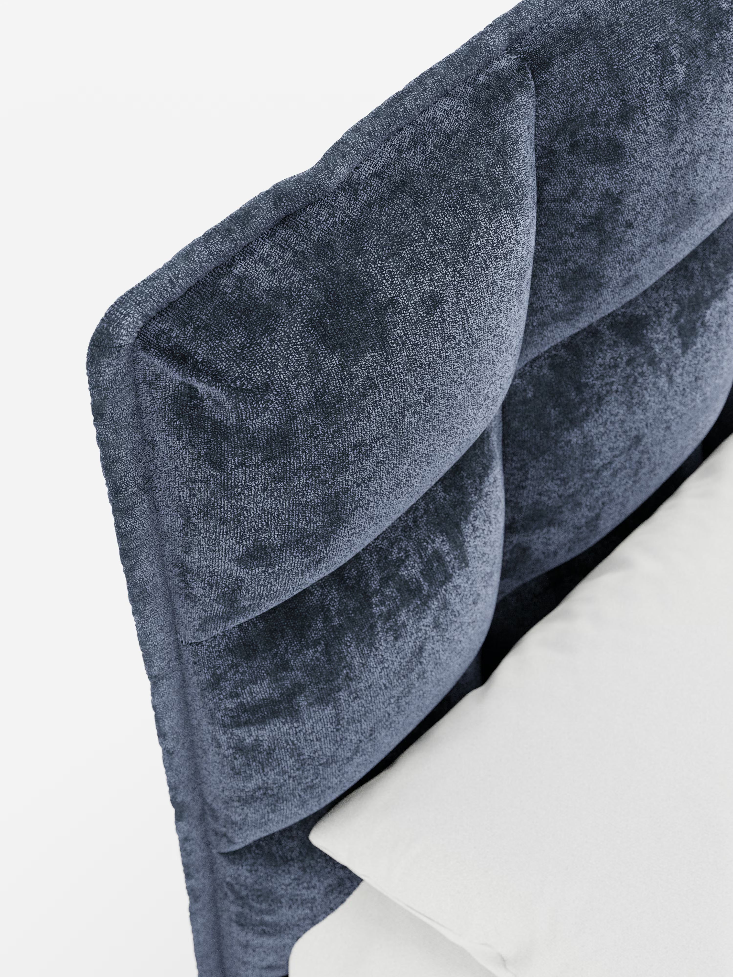 Primo piano dell'angolo del letto imbottito AURO di Letti.store, caratterizzato da un morbido rivestimento in ciniglia blu scuro certificato Oeko-Tex® e da una testiera alta, capitonné, con imbottitura a quadrati. Un cuscino bianco è appoggiato alla parte inferiore di questo elegante letto moderno con contenitore. Lo sfondo è semplice e ben illuminato.