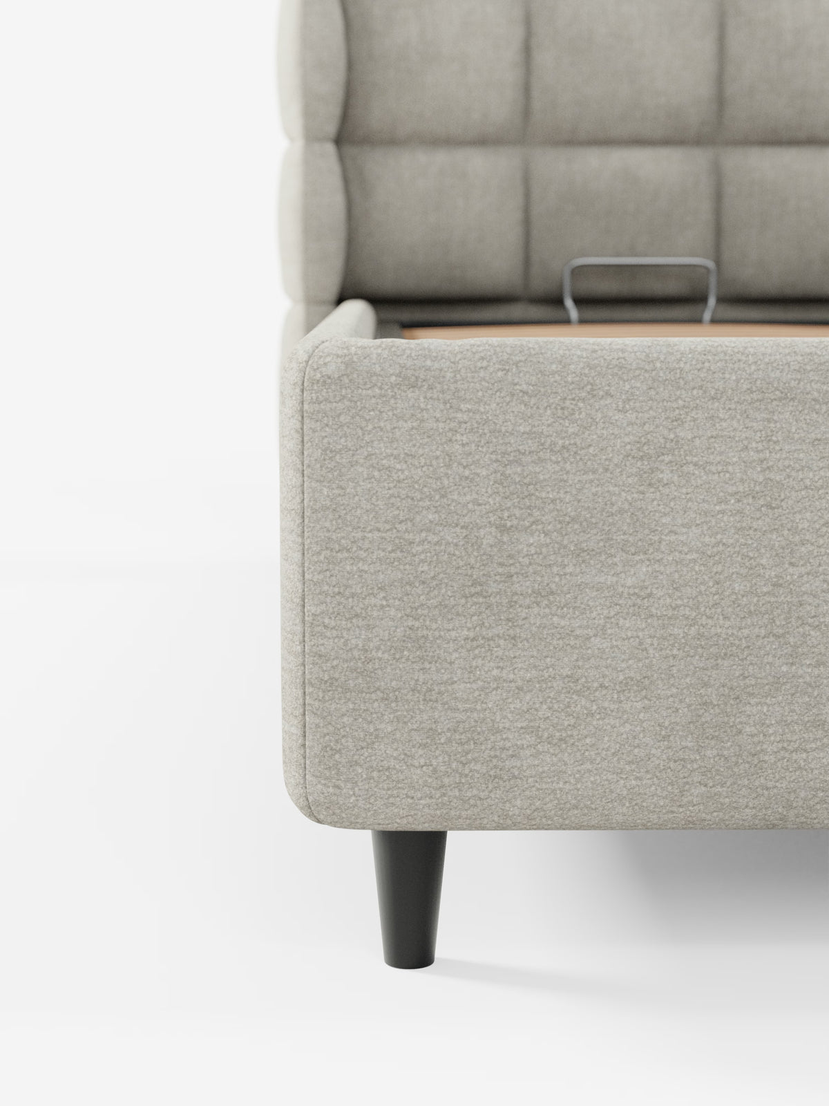 Primo piano del letto imbottito RAGNAR con vano contenitore e testiera capitonné avvolgente di Letti.store, realizzato in un elegante tessuto grigio chiaro certificato Oeko-Tex®. L'immagine mette in risalto l'angolo destro del letto, con la sua gamba affusolata nera e il pratico vano contenitore sotto il letto dotato di maniglia a vista, che lo conferma come un modello contenitore salvaspazio. Lo sfondo è bianco e spoglio, per un look pulito.
