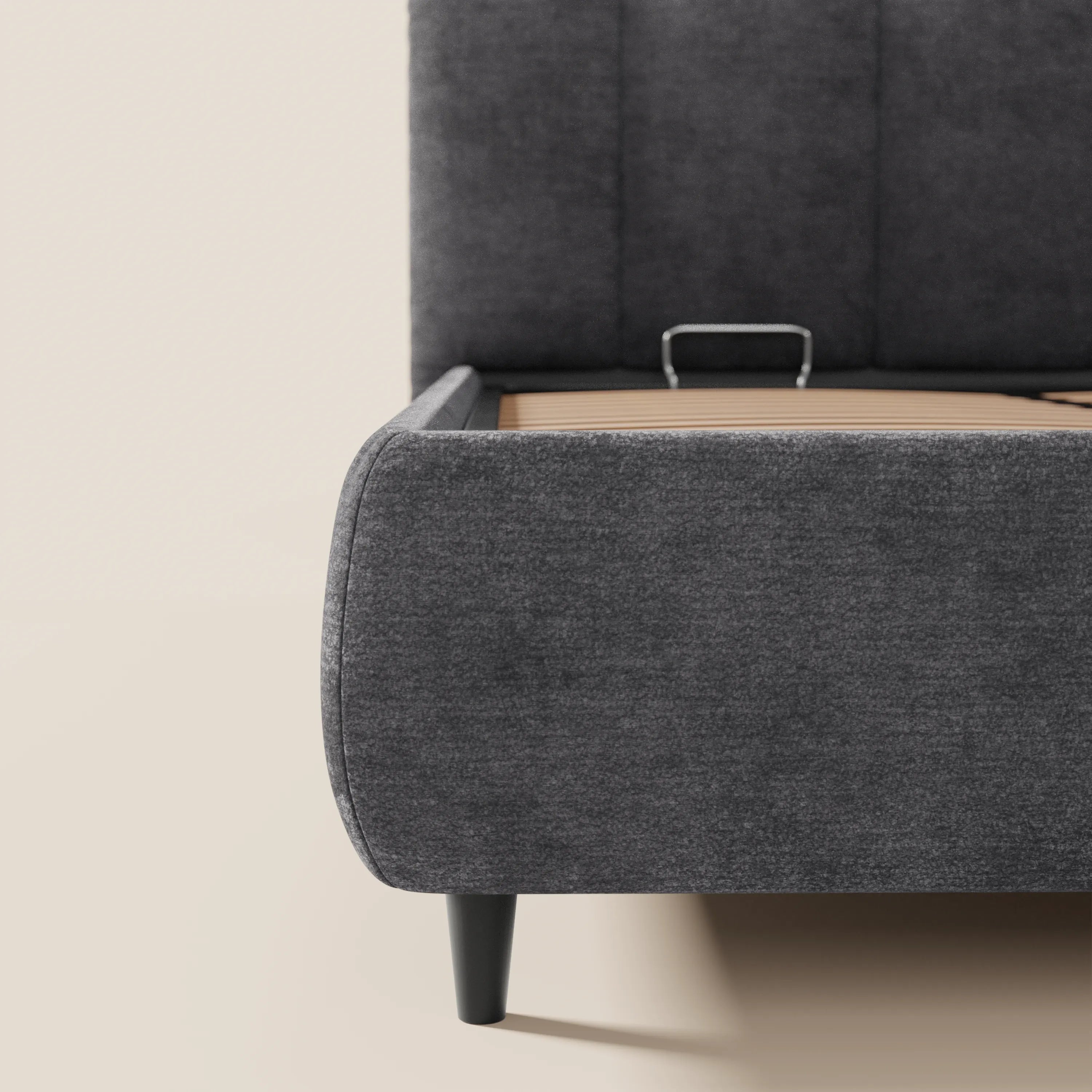 Primo piano del letto imbottito ARKAM con vano contenitore e testiera a lavorazione verticale di Letti.store, che ne mette in risalto il tessuto grigio scuro certificato Oeko-Tex® e le gambe nere, con dettagli di design in stile shabby chic su sfondo di pareti e pavimento beige chiaro.