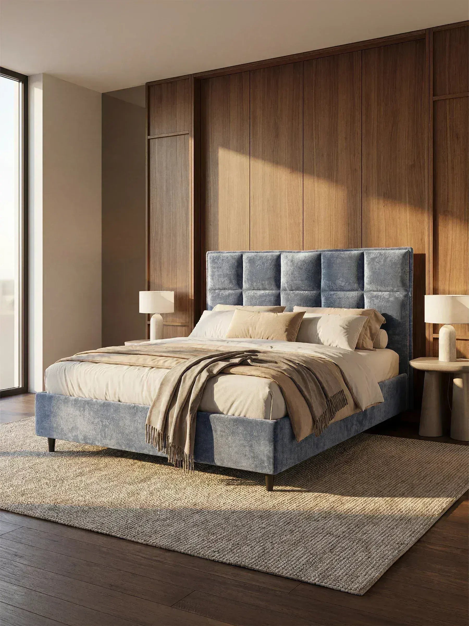 Una camera da letto moderna è arredata con il letto imbottito AURO con contenitore e testiera capitonné alta in ciniglia Oeko-Tex® di Letti.store, rivestito in velluto blu. La camera da letto è impreziosita da biancheria da letto beige, cuscini decorativi e copriletto dai toni neutri, mentre ai lati del letto si trovano due comodini rotondi con lampade bianche. Le pareti rivestite in legno e le ampie finestre garantiscono un'abbondante illuminazione naturale.