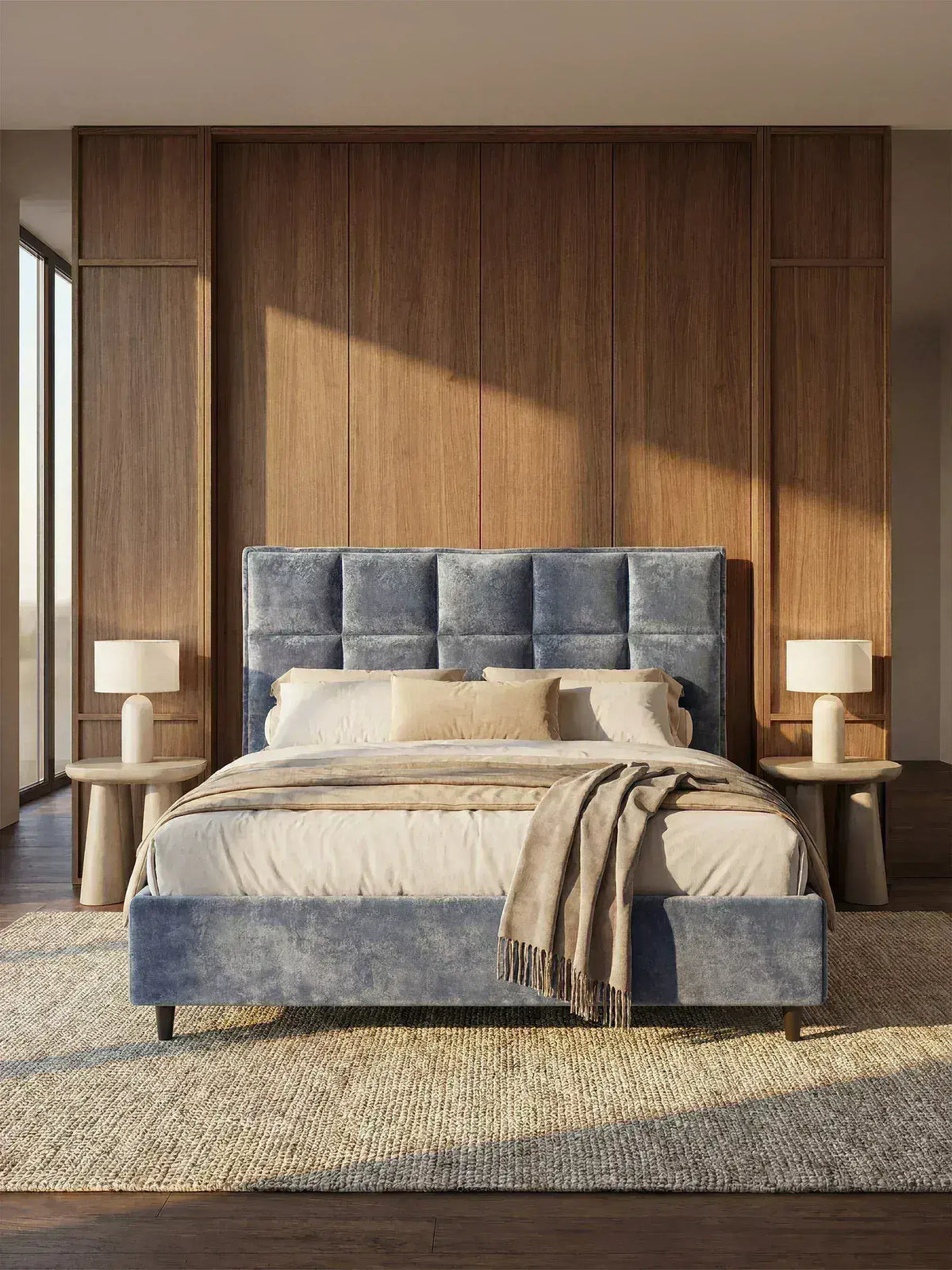 Una camera da letto moderna arredata con il letto imbottito AURO con contenitore e testiera capitonné alta in ciniglia Oeko-Tex® di Letti.store, in velluto blu, abbinato a biancheria da letto beige, due lampade beige sui comodini in legno e un tappeto dalla trama rigata. La parete dietro al letto è impreziosita da un rivestimento verticale in legno, mentre la luce del sole filtra da una finestra sulla sinistra.