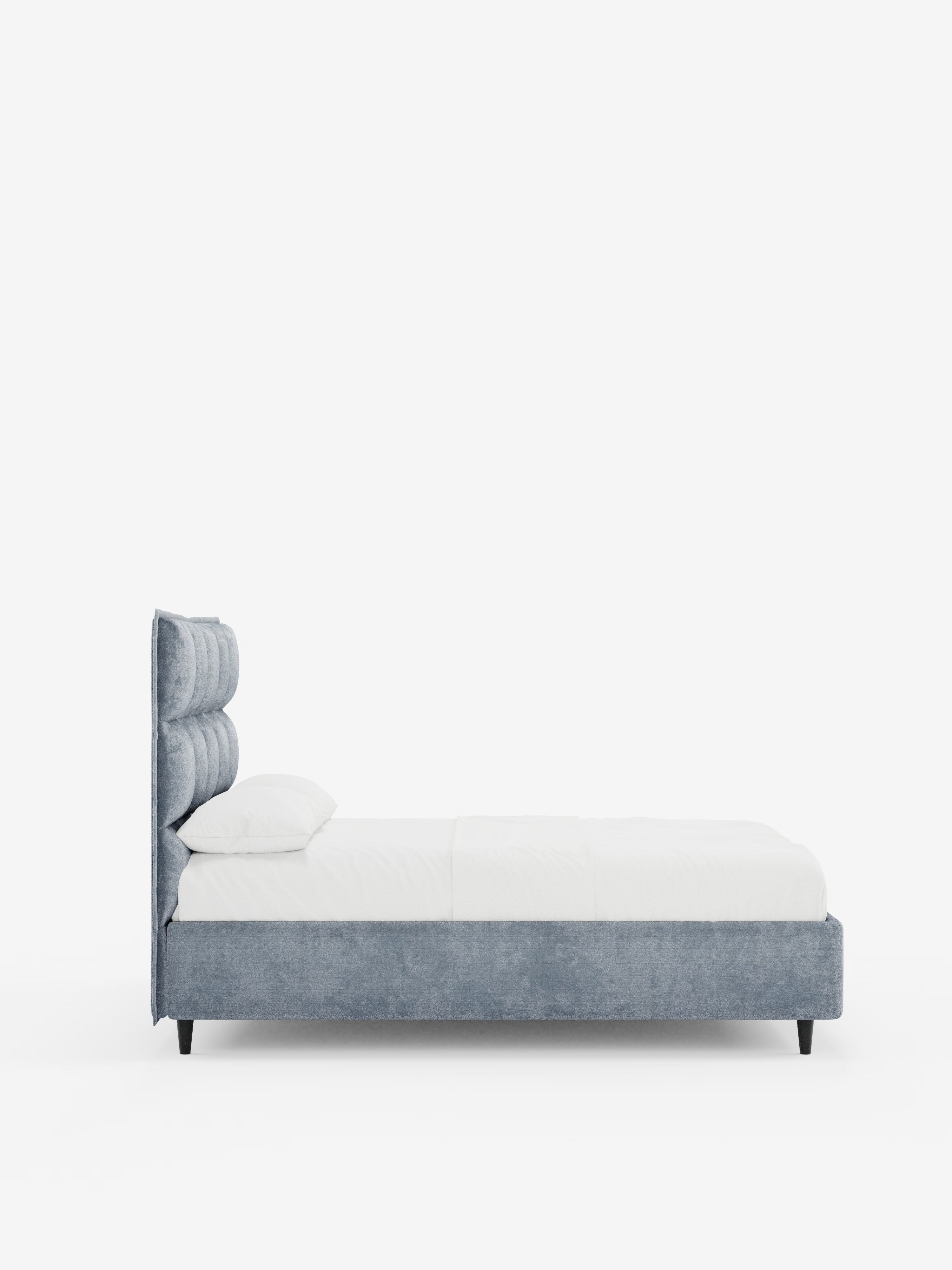 Vista laterale del letto imbottito AURO con vano contenitore di Letti.store, caratterizzato da un’alta testiera capitonné in tessuto chenille azzurro certificato Oeko-Tex®. Il letto è abbinato a biancheria da letto bianca e due cuscini, su uno sfondo bianco a tinta unita che ne esalta il design minimalista e la morbida trama.