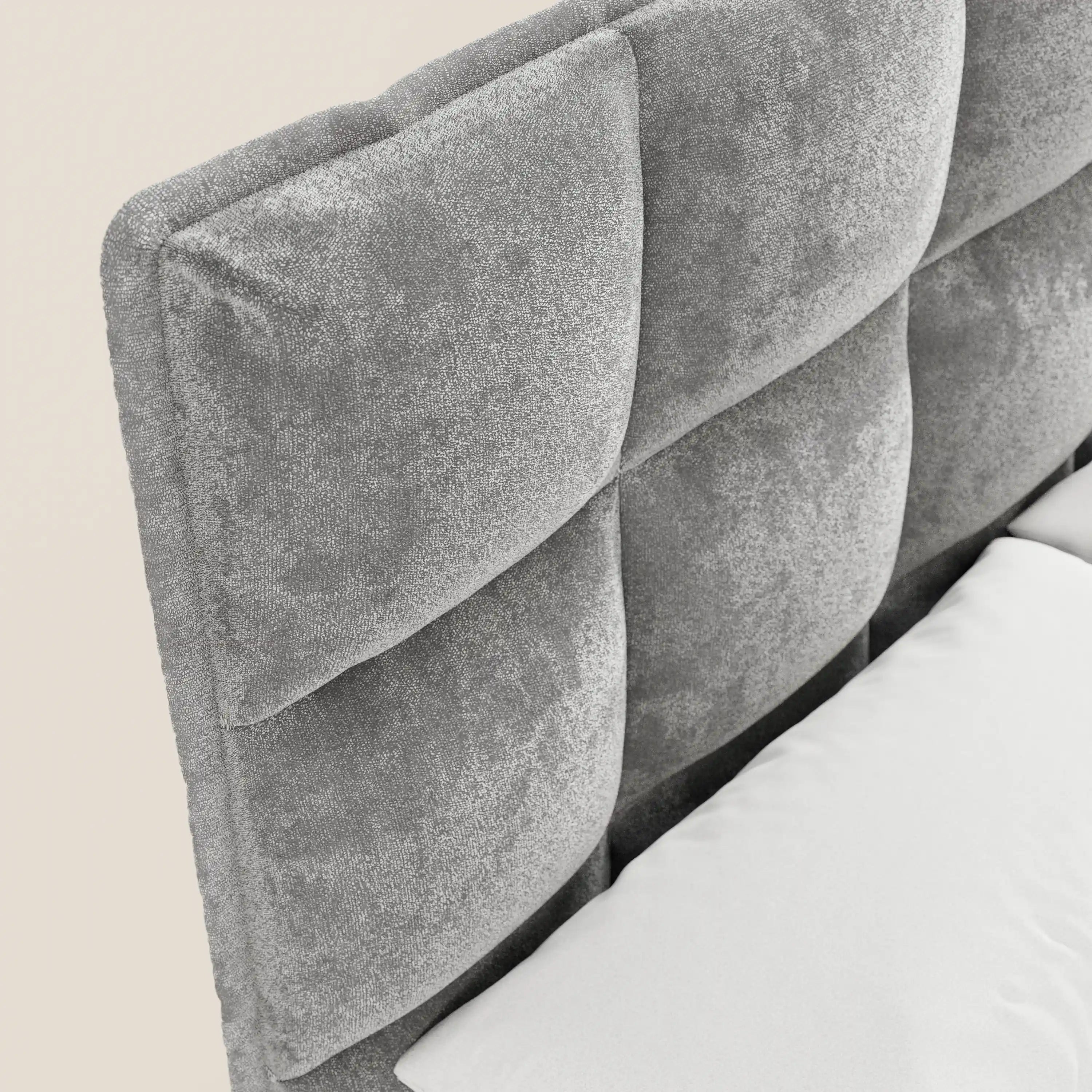 Primo piano del letto imbottito AURO di Letti.store, che mette in risalto la sua alta testiera capitonné in morbida ciniglia certificata Oeko-Tex®. Le ampie sezioni imbottite di forma quadrata e un cuscino bianco risaltano sullo sfondo neutro.