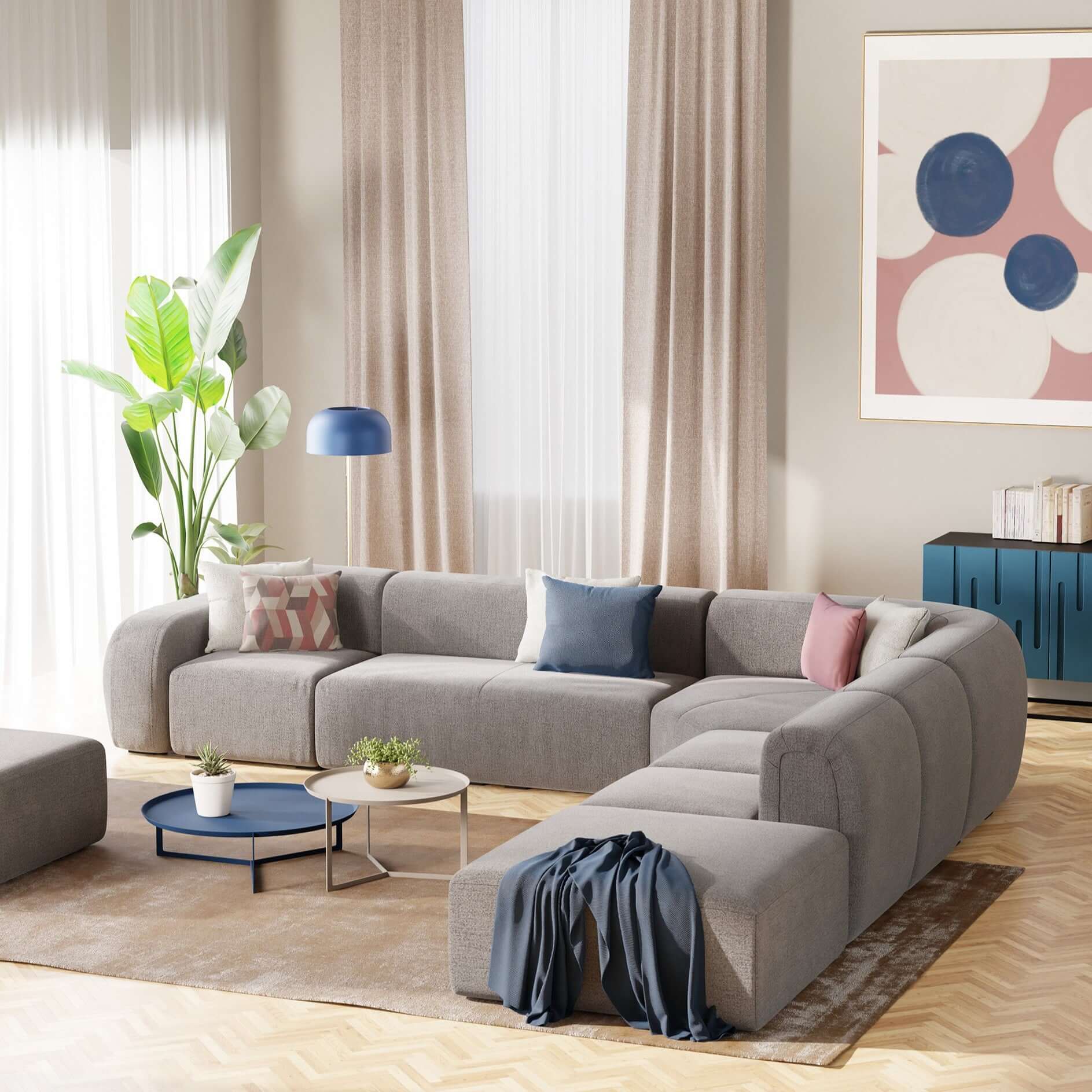 Un soggiorno moderno è caratterizzato dal divano BonBon angolare componibile e modulare in tessuto smacchiabile T23 grigio di Divani.Store, con cuscini azzurri e rosa, giaciglio blu, tavolini rotondi, piante, opere d'arte astratta a parete, credenza blu su un tappeto beige e pavimento in legno.