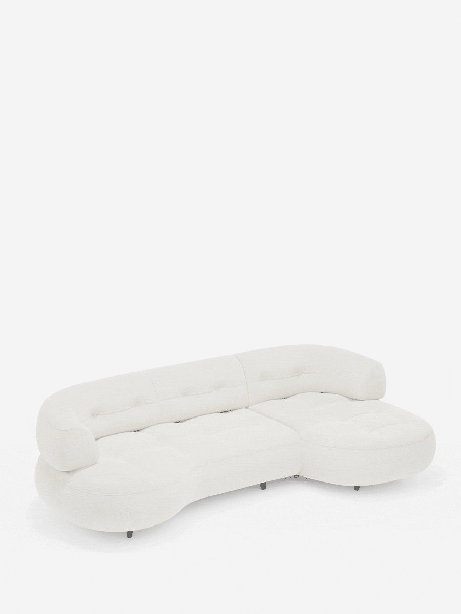 Un divano angolare Croissant color crema, dalla forma a fagiolo, con schienali scorrevoli rivestiti in morbido tessuto effetto cotone smacchiabile T33, dal design modulare sinuoso, cuscini arrotondati e piedini scuri dal profilo ribassato.