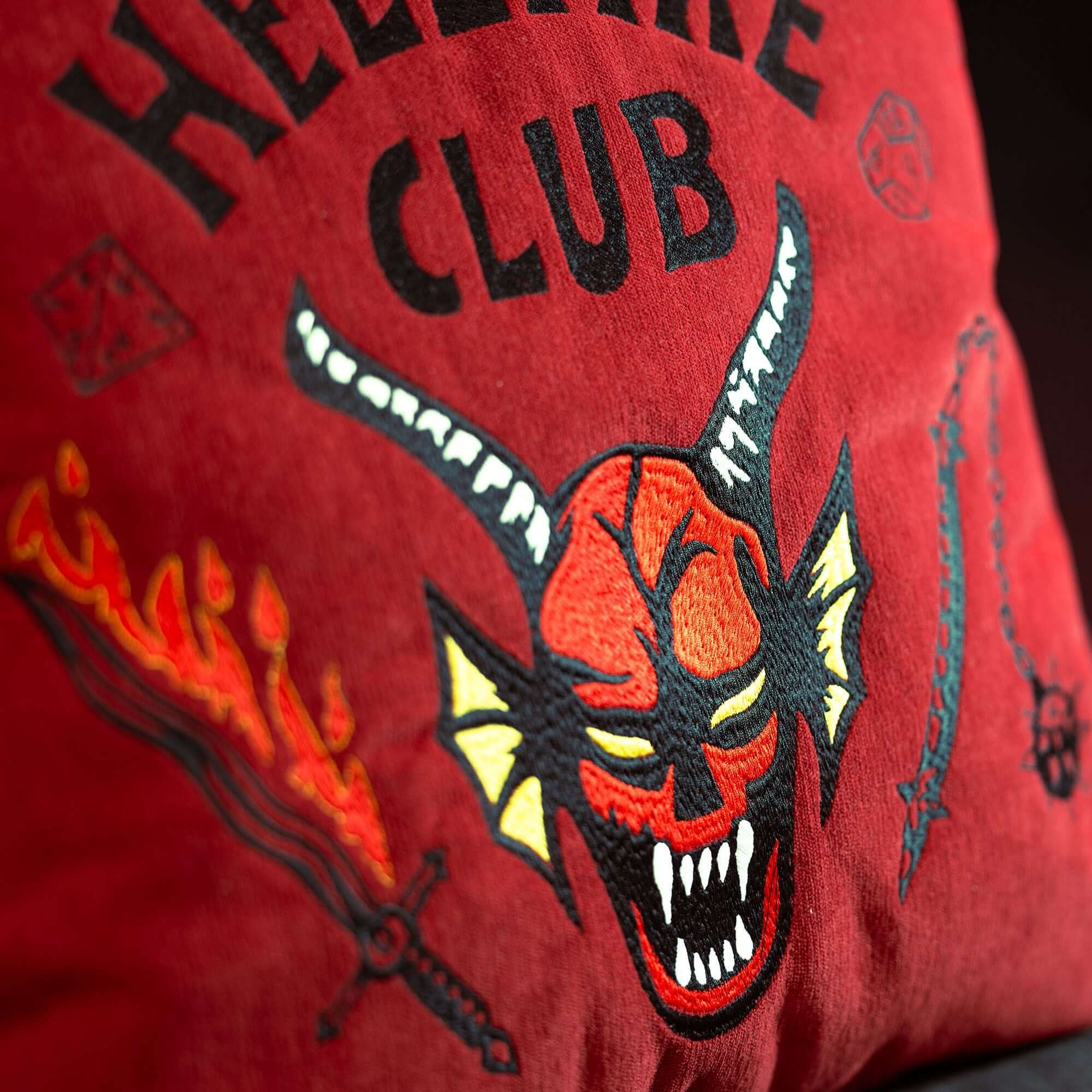 Primo piano del Cuscino Hellfire club Stranger Things Limited Edition di Divani.Store: rosso 45x45 cm, tessuto antimacchia, faccia di demone ricamata con "HELLFIRE CLUB", dadi, spada infuocata e fiocina chiodata.