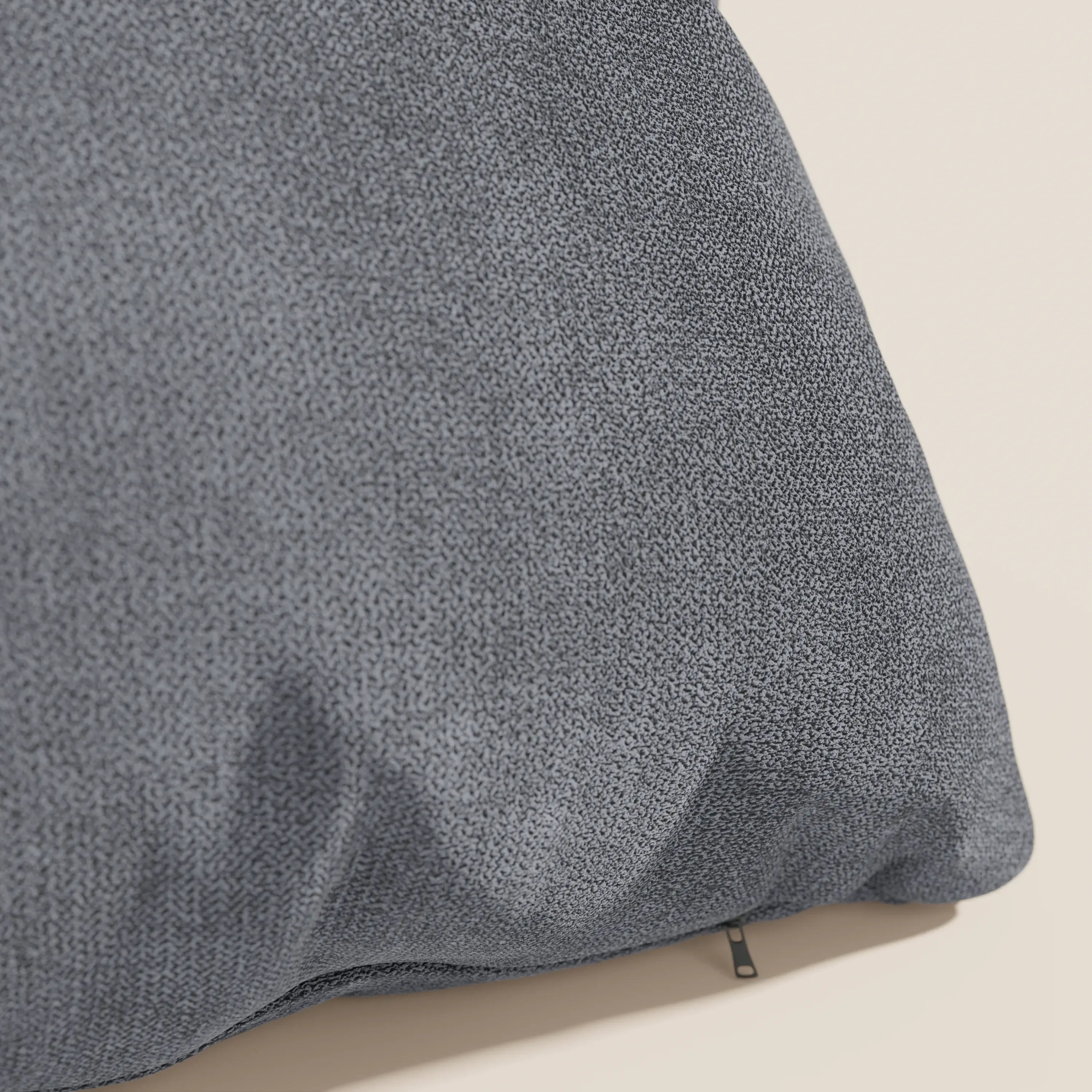 Un primo piano del cuscino Destiny di Divani.Store in tessuto morbido impermeabile T02 grigio mostra la sua superficie morbida e sottilmente strutturata con pieghe e una piccola cerniera nera vicino al fondo, su uno sfondo beige chiaro.
