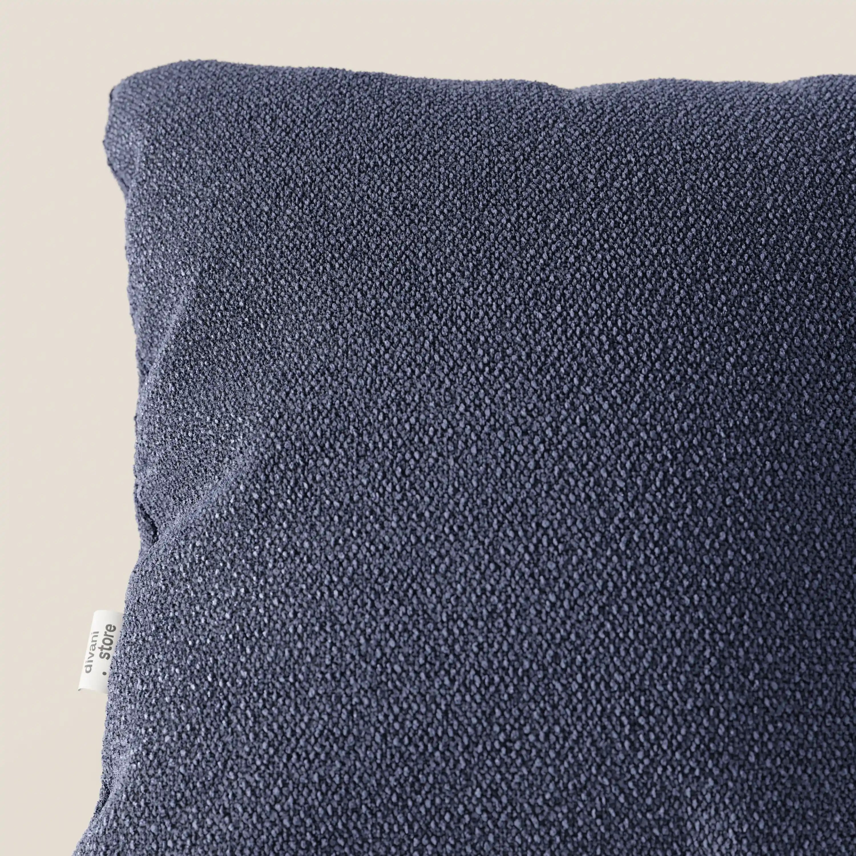 Primo piano dell'angolo superiore sinistro di un cuscino Lambada blu scuro di Divani.Store, realizzato in tessuto smacchiabile e impermeabile tipo cotone naturale, con una piccola etichetta bianca del marchio e una texture leggermente sassosa su uno sfondo chiaro.