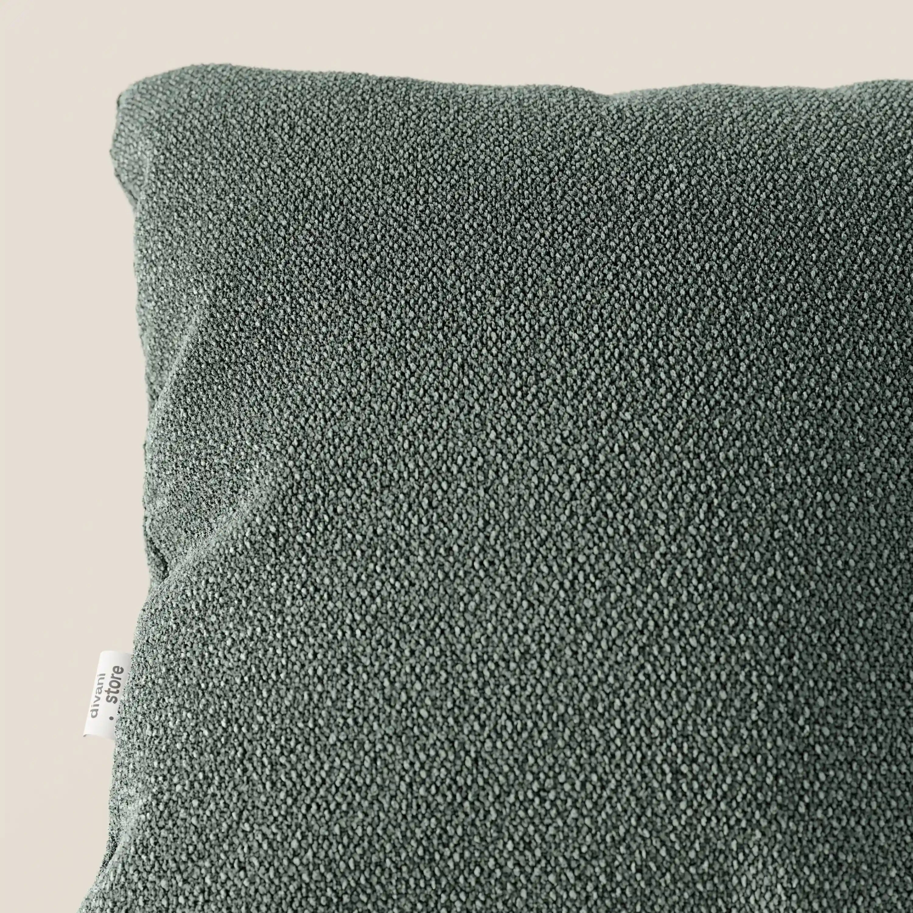 Primo piano del cuscino Lambada di Divani.Store, di colore verde scuro, realizzato in tessuto fresco, antimacchia e impermeabile tipo cotone (T33), appoggiato su una superficie beige chiaro. Sul bordo sinistro compare una piccola etichetta bianca, visibile solo in parte.
