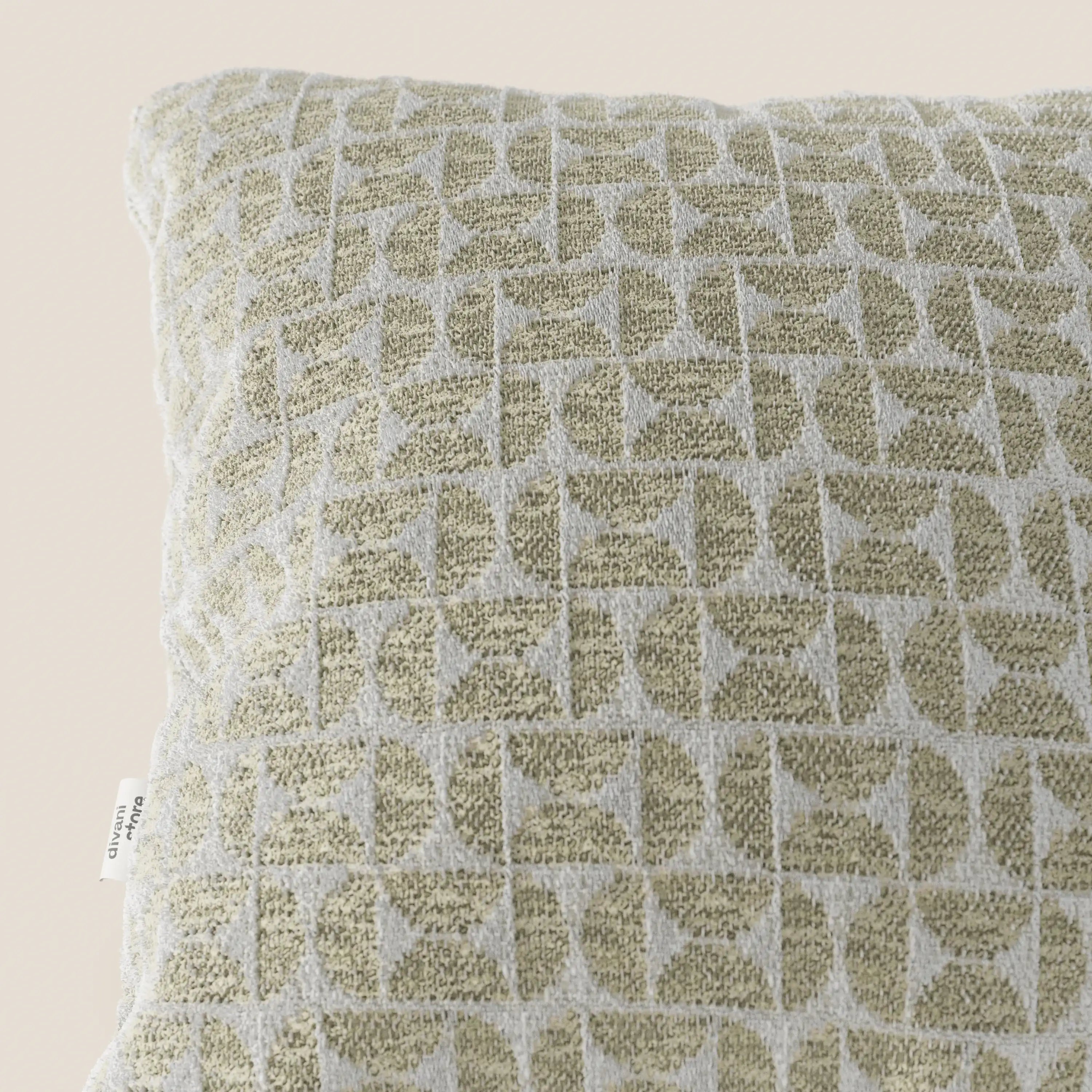 Un primo piano del cuscino "Naxos Cuscino Decoro 45x45 in Lino" di Divani.Store mette in risalto il suo elegante motivo geometrico nei toni del verde chiaro e del bianco sporco, con un disegno in rilievo composto da semicerchi e quadrati, oltre a un'etichetta in tessuto bianca sul bordo sinistro.