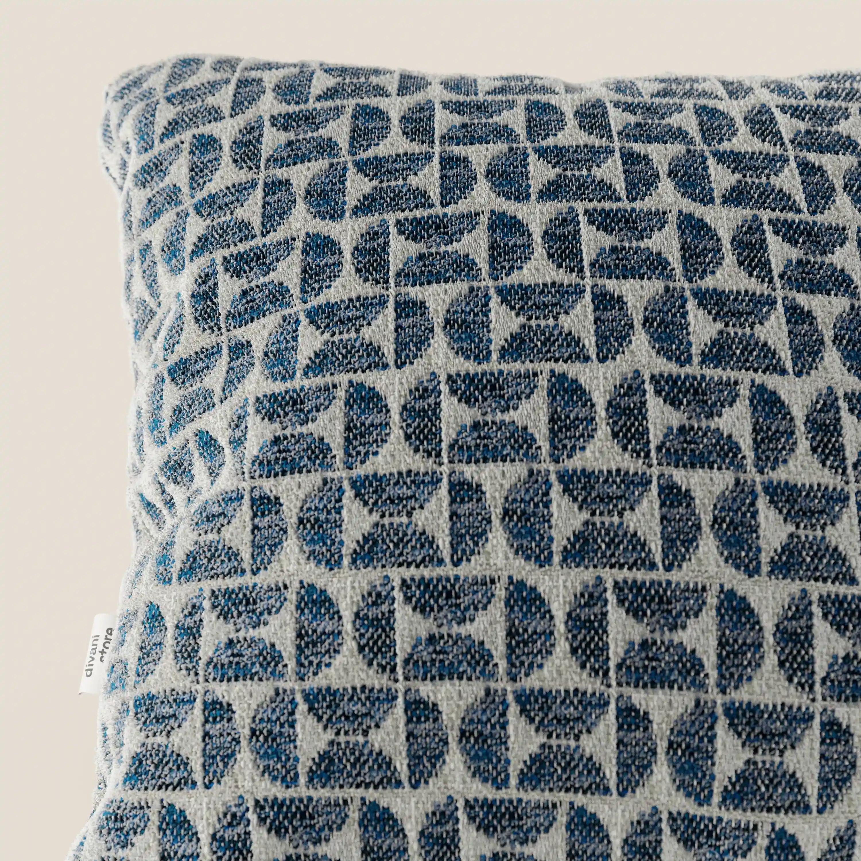 Primo piano del cuscino Naxos Decoro 45x45 in lino di Divani.Store, caratterizzato da un elegante motivo geometrico blu su lino grigio chiaro. A sinistra è visibile una piccola etichetta bianca; lo sfondo è a tinta unita e di colore chiaro.