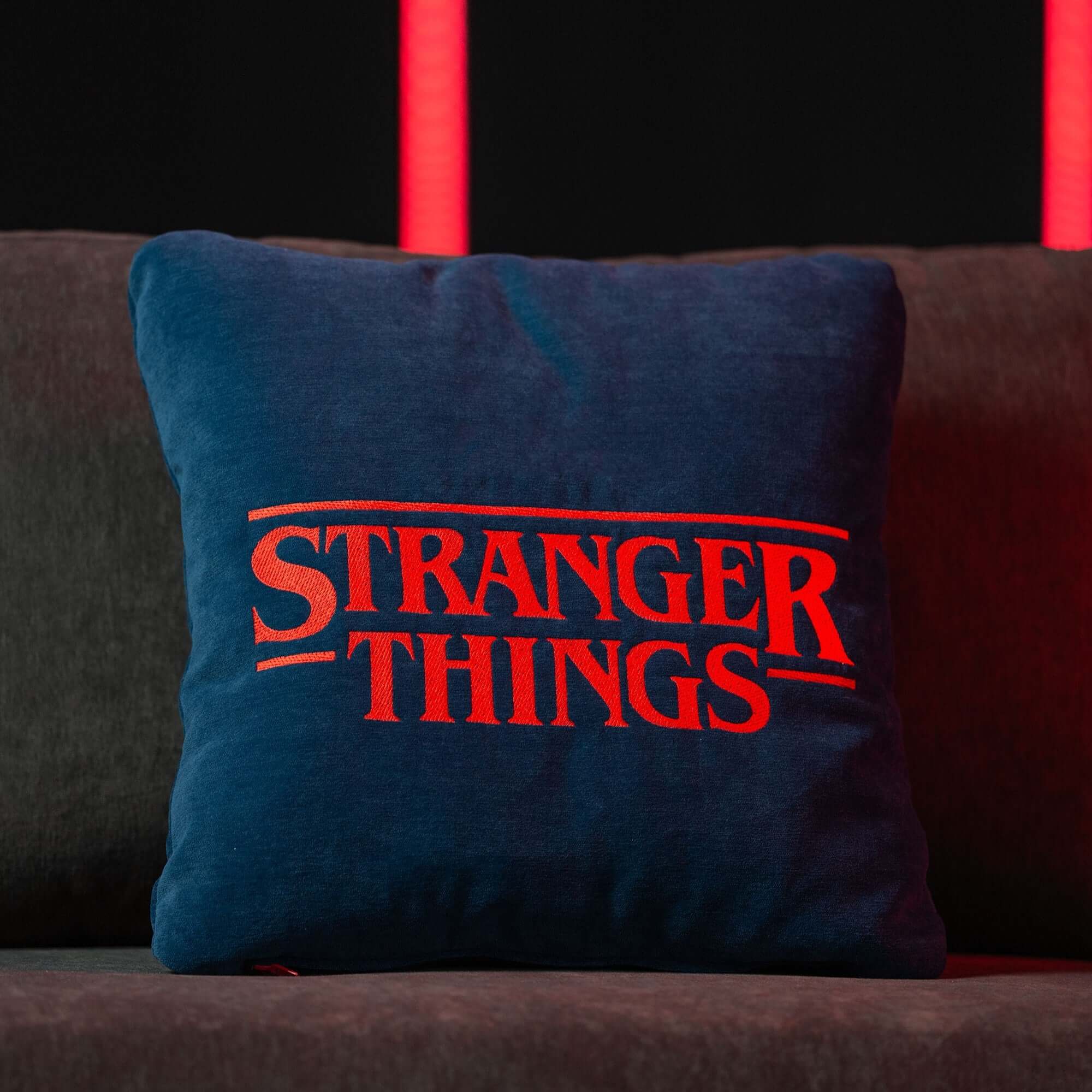 Un Cuscino Stranger Things Limited Edition con Ricamo 45x45 blu di Divani.Store si trova su un divano grigio scuro, illuminato da luci rosse verticali su sfondo nero: un accessorio d'arredo accattivante e di grande effetto.