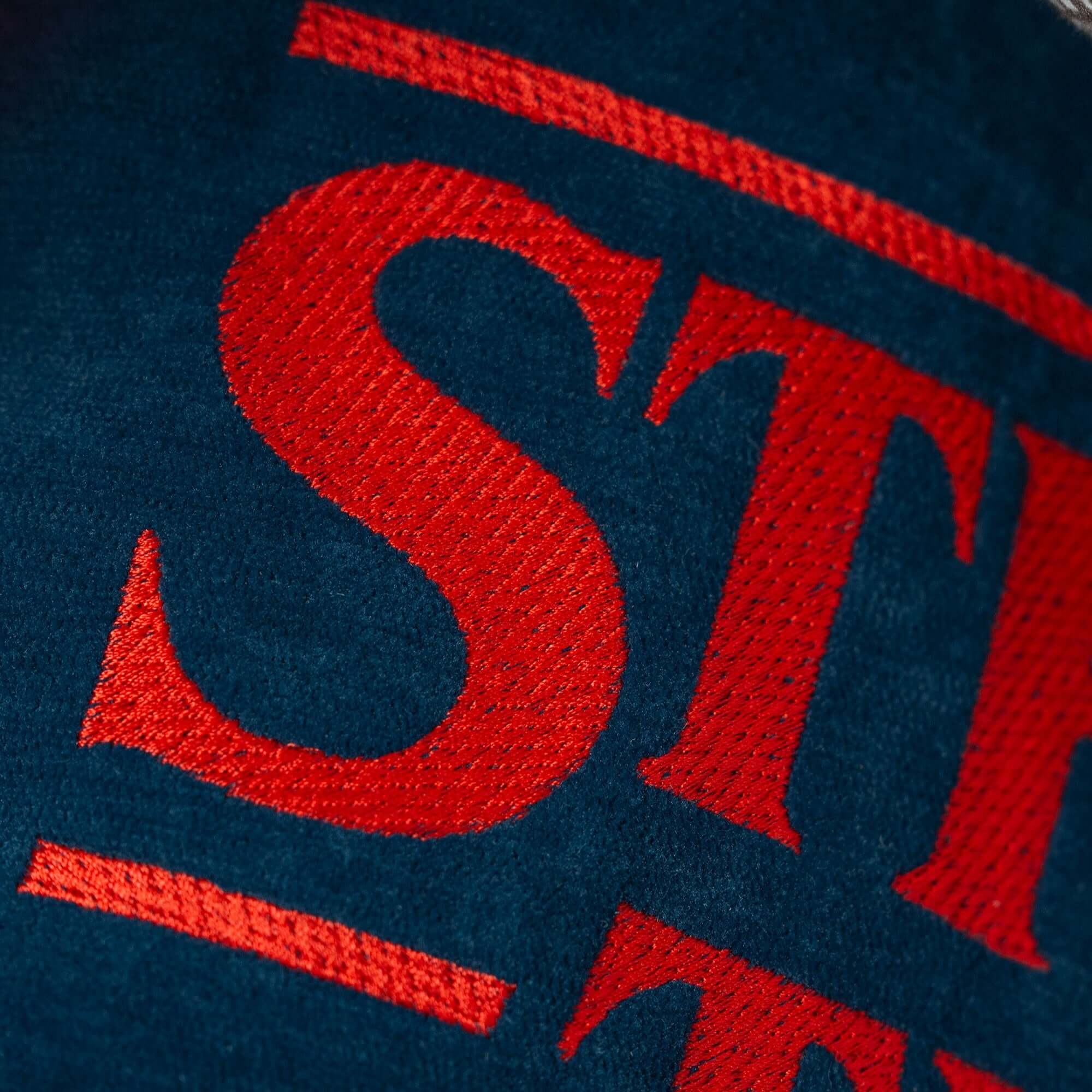 Primo piano della scritta "STI" ricamata in rosso su tessuto blu scuro antimacchia del Cuscino Stranger Things Limited Edition con Ricamo 45x45 di Divani.Store. Sono visibili le cuciture dettagliate e parte della sottolineatura; si vede solo una parte del logo.