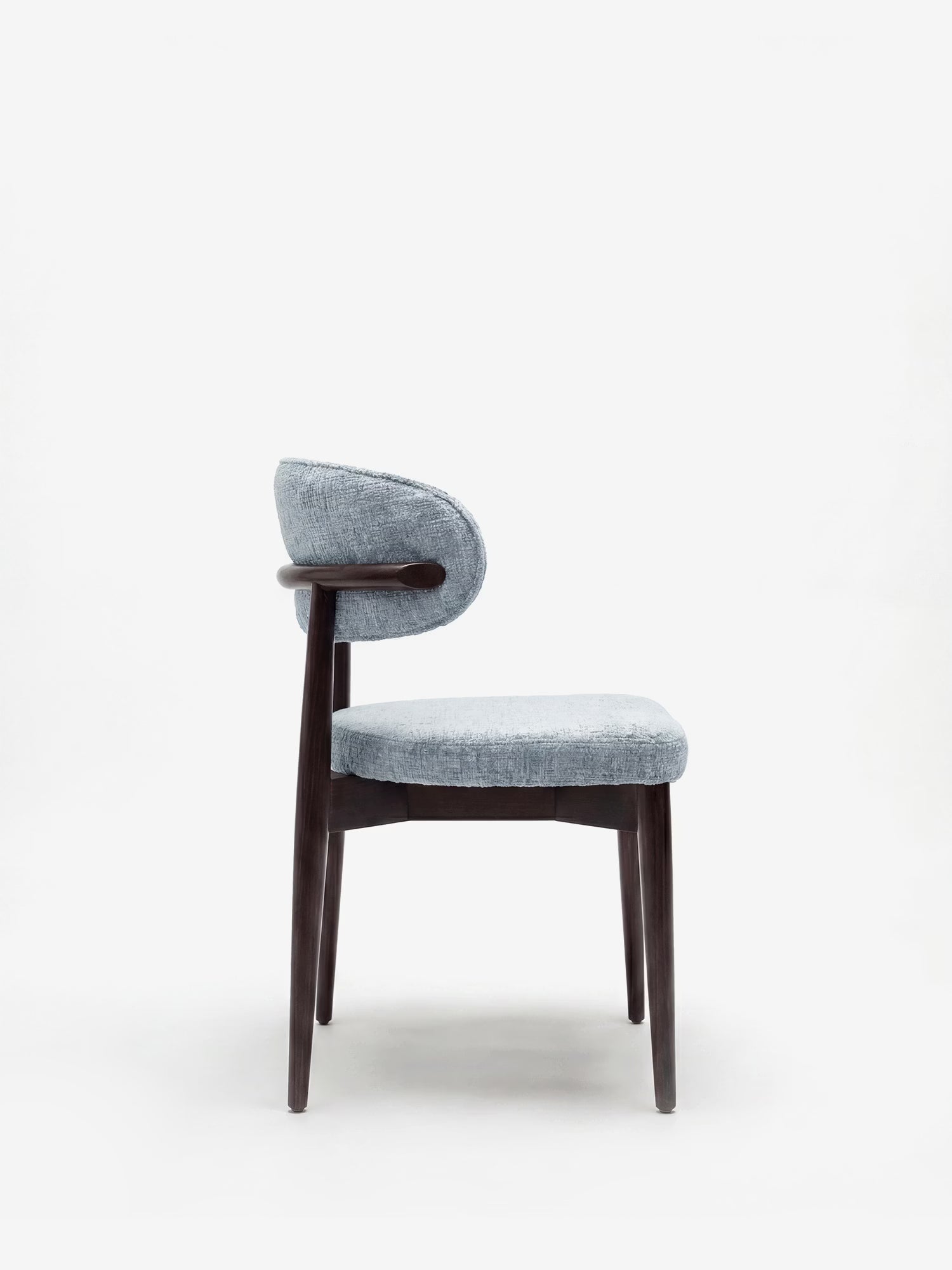 La sedia di design Dafne di Divani.Store presenta una seduta arrotondata in ciniglia azzurra certificata OEKO-TEX® e uno schienale curvo, sostenuti da quattro gambe in legno massello di wengé. Fotografata di profilo su uno sfondo bianco a tinta unita.