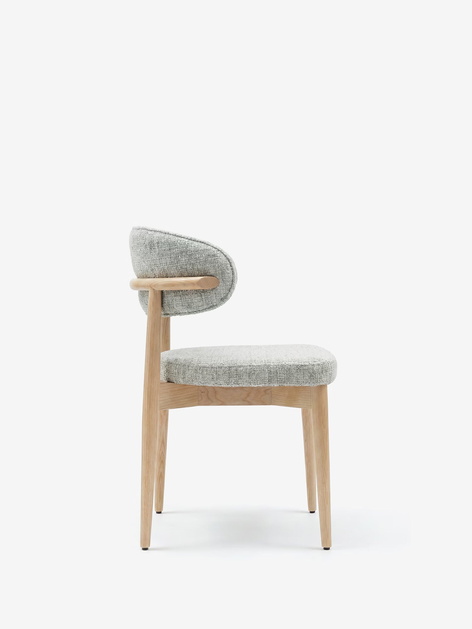 La sedia imbottita Dafne di Divani.Store presenta gambe in rovere massello naturale, uno schienale imbottito dalla linea curva e una seduta imbottita in ciniglia grigio chiaro certificata OEKO-TEX®, qui raffigurata di profilo su sfondo bianco.