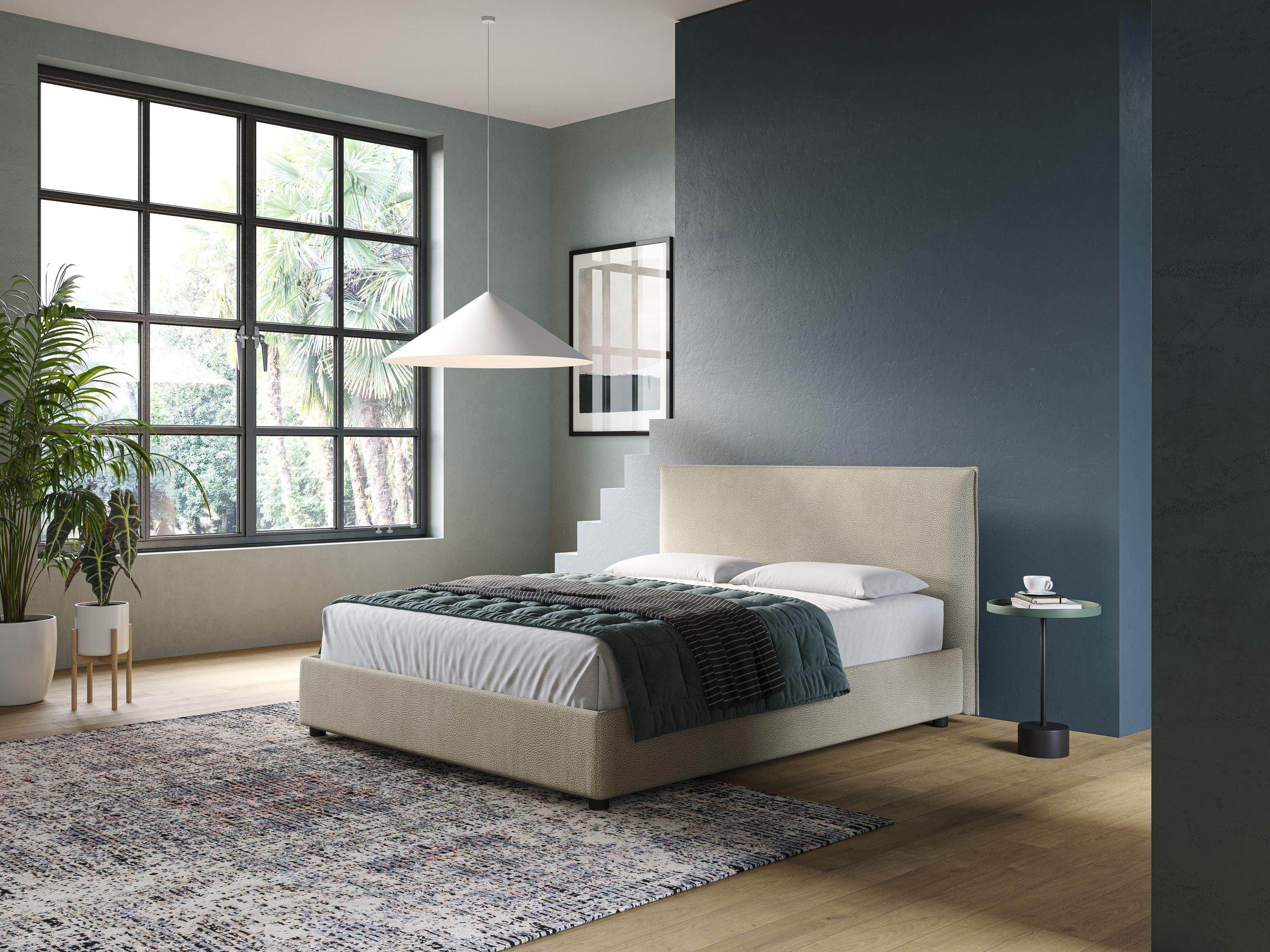 Una camera da letto moderna con un'ampia finestra, piante verdi e una parete d'accento blu è caratterizzata dal letto contenitore Nina dal design minimal in microfibra effetto pelle smacchiabile panna di Letti.store. Un tavolino rotondo contiene una tazza e un piattino; una lampada a sospensione bianca è appesa sopra un tappeto.