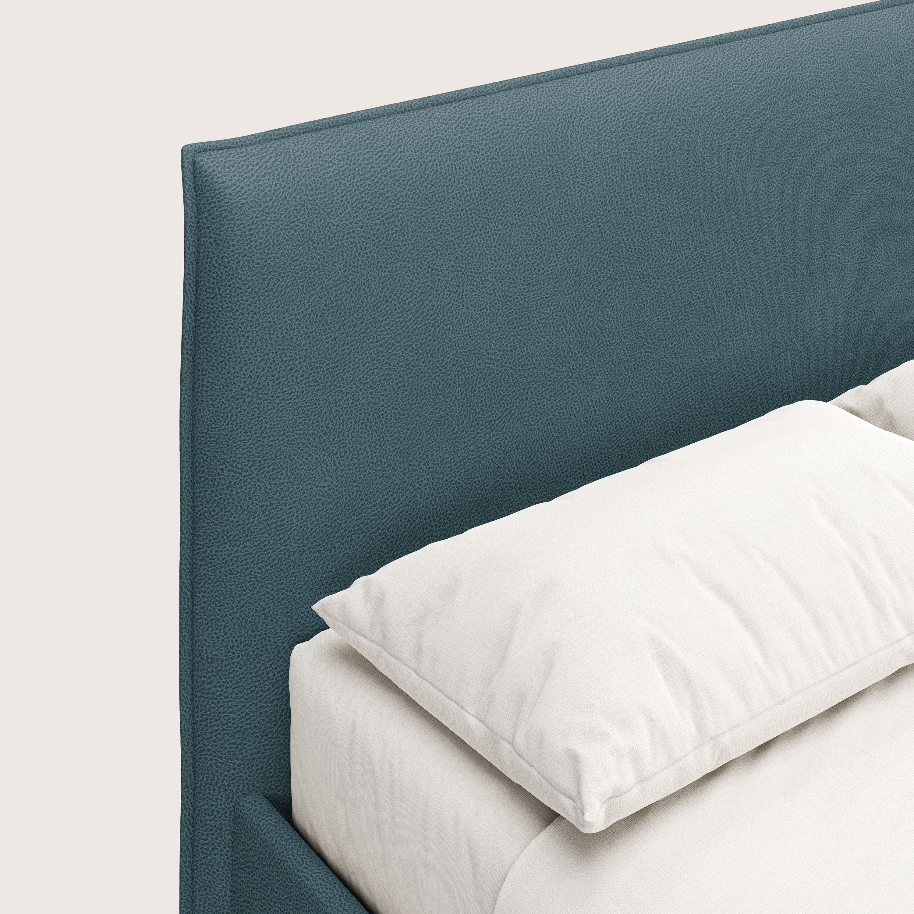 Primo piano del letto contenitore Nina di Letti.store, caratterizzato da un design minimale con rivestimento in microfibra di petrolio testurizzata e antimacchia effetto pelle, ben realizzato con lenzuola bianche e un cuscino in peluche su uno sfondo chiaro.