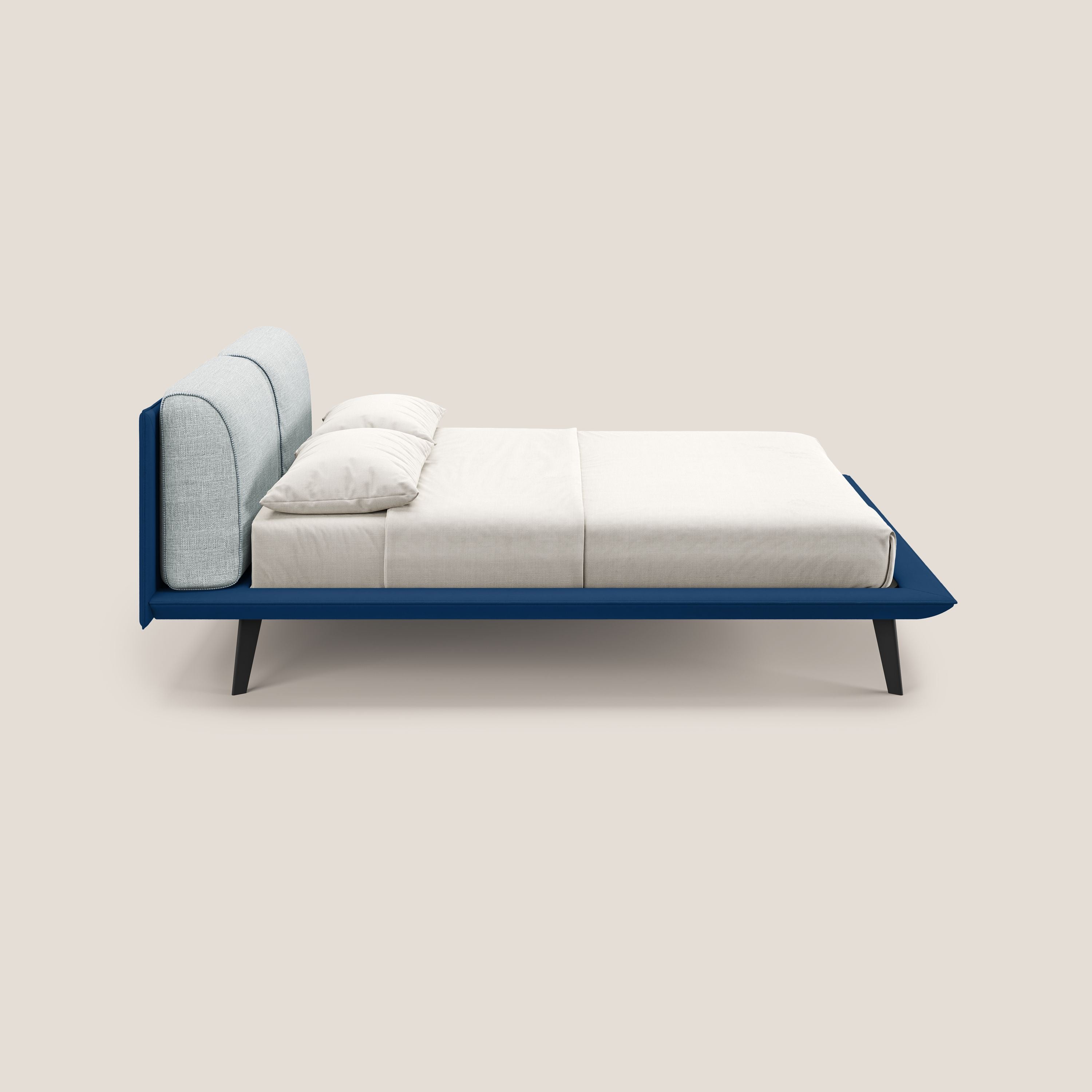 Il letto di design Petra di Letti.store è caratterizzato da un tessuto impermeabile bicolore blu-ghiaccio, cuscini in piuma e gambe angolate in mogano. Viene mostrato ben realizzato con biancheria da letto bianca in un profilo laterale su sfondo beige.