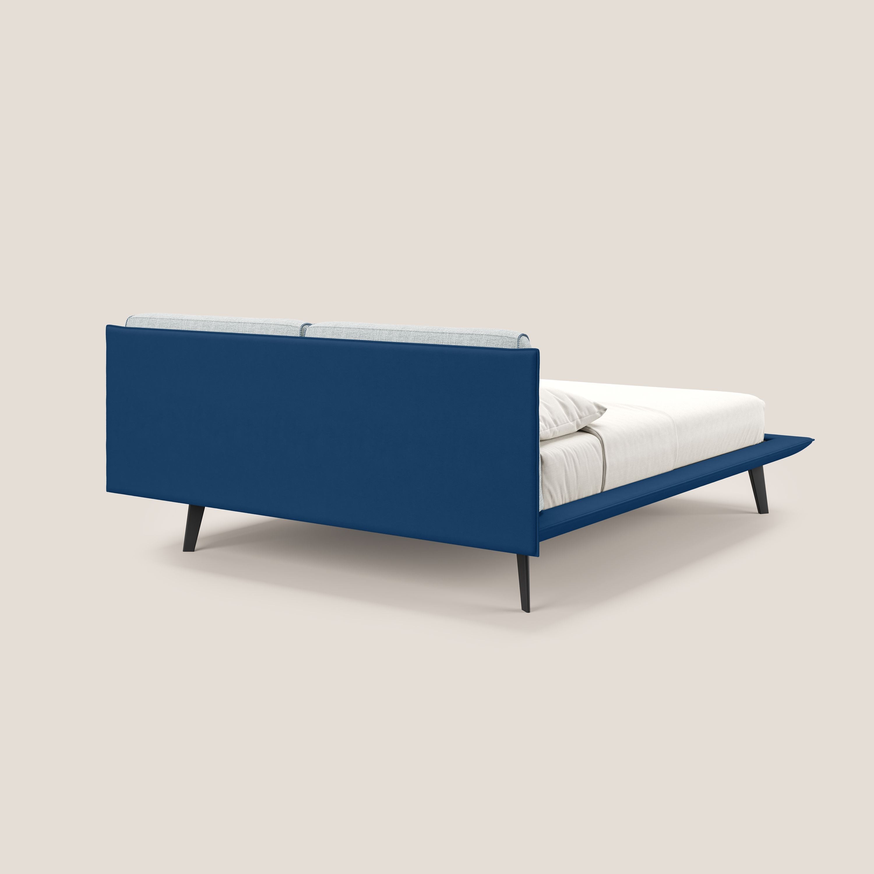 Il letto di design Petra di Letti.store è caratterizzato dal tessuto impermeabile bicolore T11 blu e T06 ghiaccio, dai cuscini in piuma e dai piedini in mogano. Il letto è presentato con biancheria bianca e due cuscini grigio chiaro su sfondo beige, visto dall'angolo posteriore sinistro.