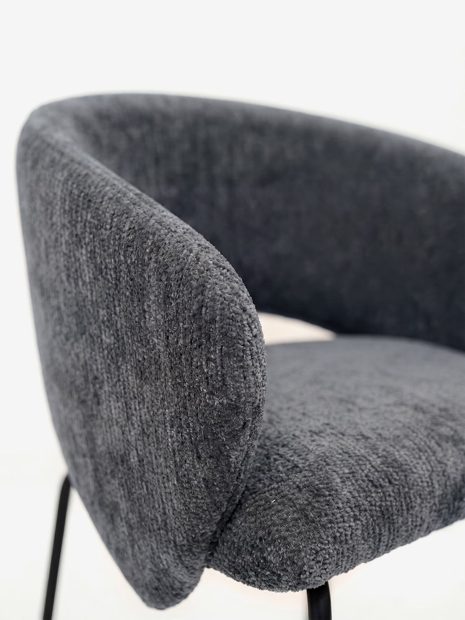 Primo piano della sedia imbottita con braccioli Fosca, un modello di design realizzato in morbido tessuto certificato OEKO-TEX® da Divani.Store, caratterizzata da uno schienale arrotondato e da sottili gambe in metallo nero, su uno sfondo bianco a tinta unita.