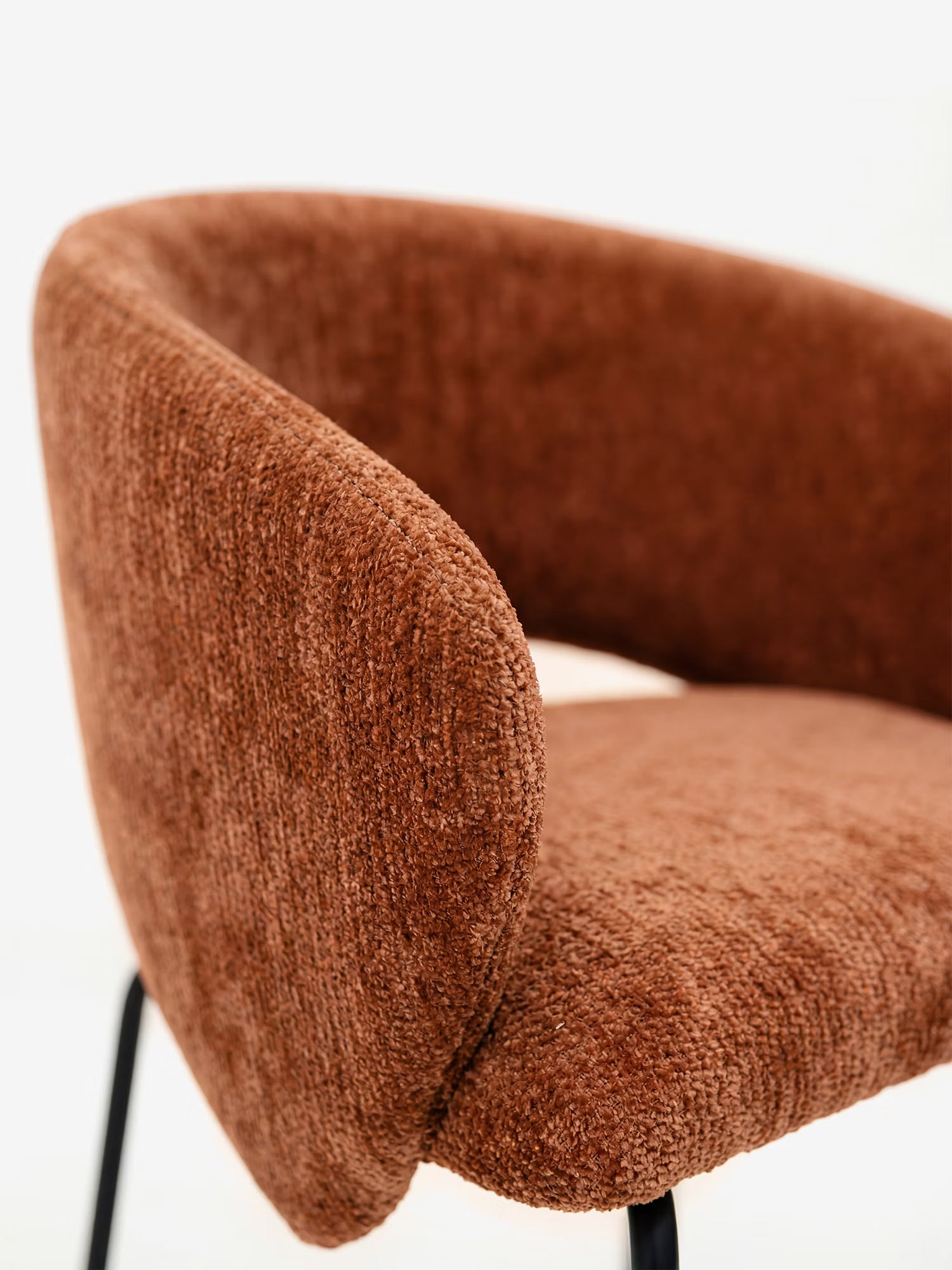 Primo piano della sedia imbottita Fosca di Divani.Store, caratterizzata da un morbido tessuto marrone certificato OEKO-TEX®, uno schienale e braccioli curvi e sottili gambe in metallo nero: il suo design moderno risalta su uno sfondo bianco a tinta unita.