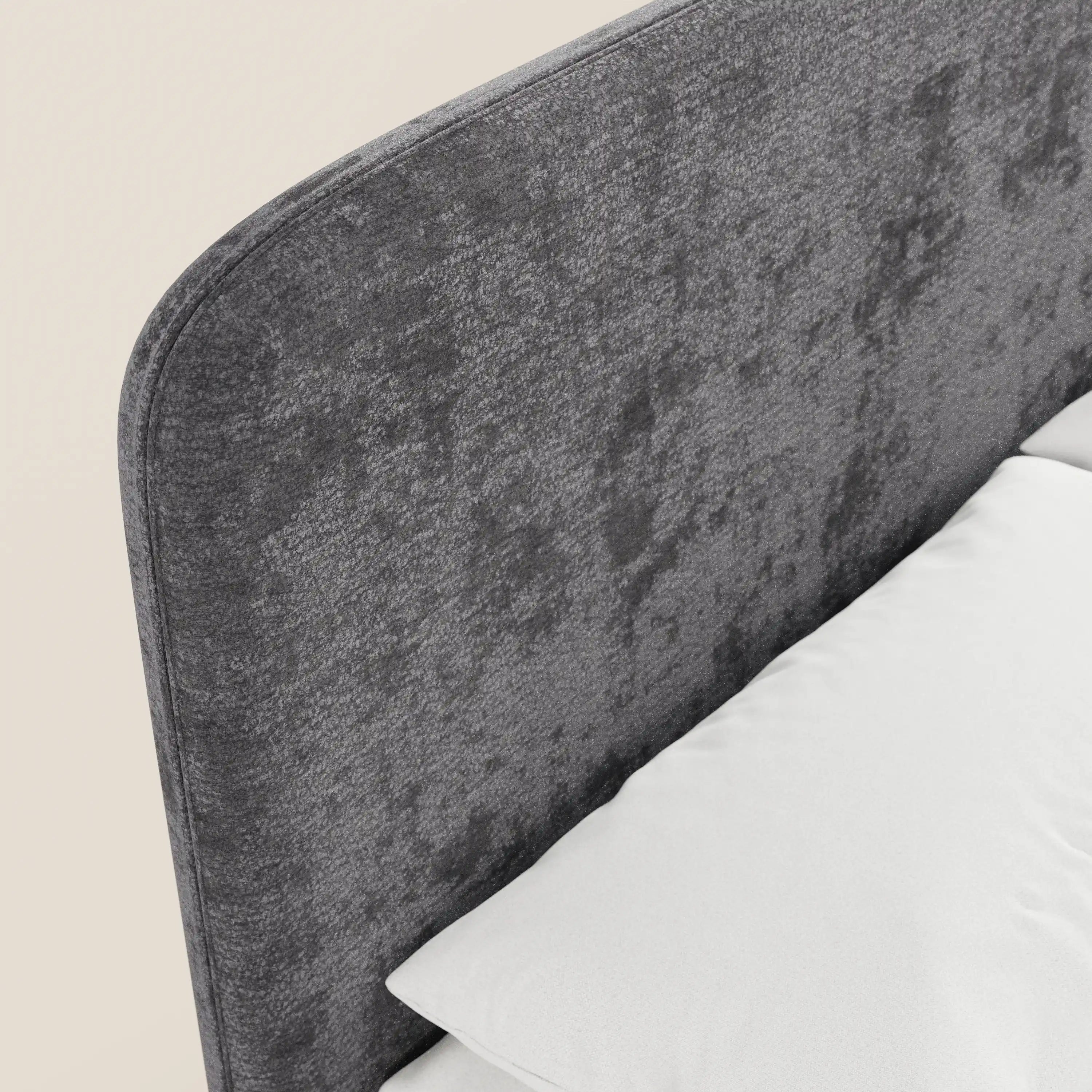 Primo piano del letto imbottito KAELOS Letto Minimal di Letti.store: testiera arrotondata rivestita in ciniglia grigia, gambe alte, vano contenitore, raffigurato accanto a un cuscino bianco e a un materasso su uno sfondo beige — ideale per una camera da letto moderna.