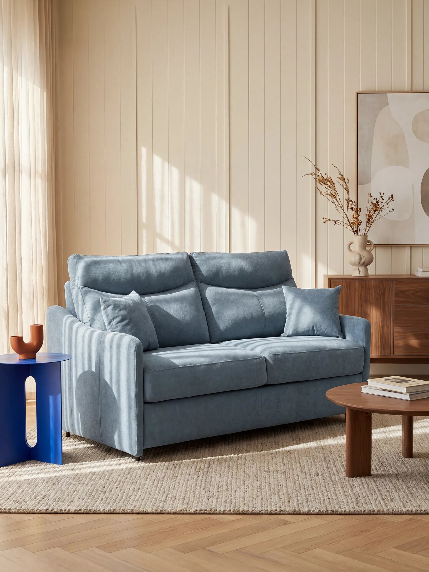 Il divano letto Lodovico a 3 posti con materasso ALTO 20 cm in tessuto smacchiabile certificato Oeko-Tex di Divani.Store, rivestito in tessuto azzurro, è disposto con due cuscini su un tappeto beige a trama ruvida. La stanza inondata di luce, con pareti rivestite di pannelli color crema, presenta anche un tavolino rotondo in legno, un piccolo tavolino blu su cui è posato un vaso marrone e una credenza in legno decorata con opere d’arte astratta e piante essiccate.