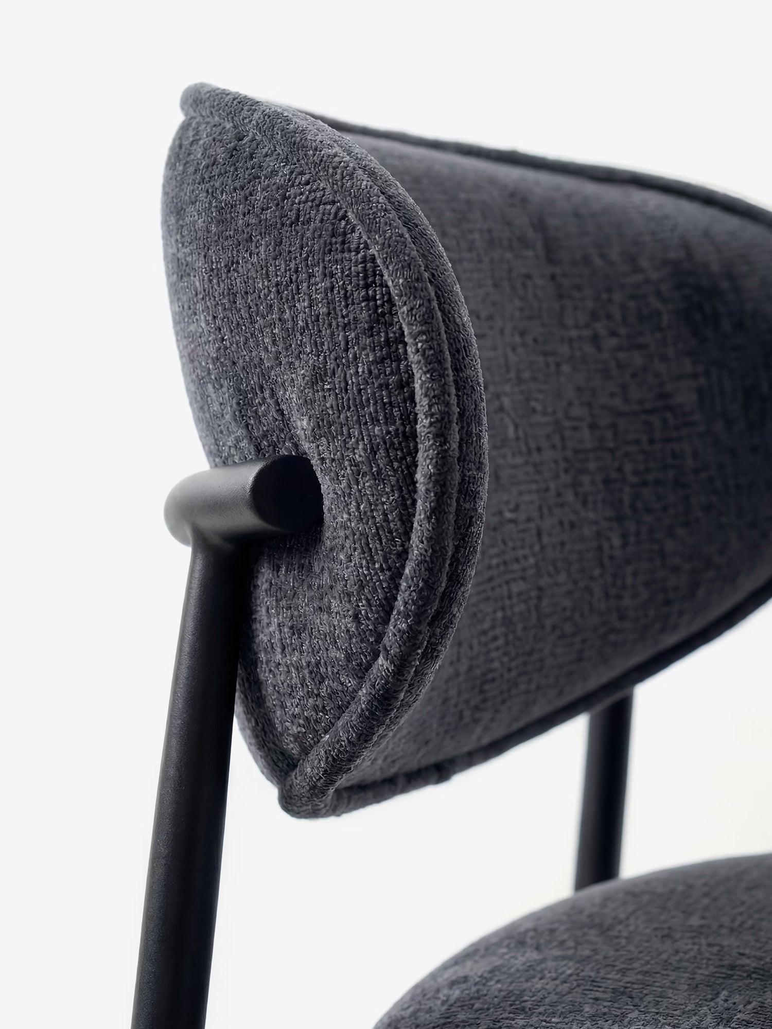 Primo piano della Luce Sedia, imbottita in morbido tessuto ciniglia di Divani.Store: presenta uno schienale curvo e imbottito in tessuto grigio scuro testurizzato e una struttura in metallo nero opaco dalle linee minimaliste su sfondo bianco.