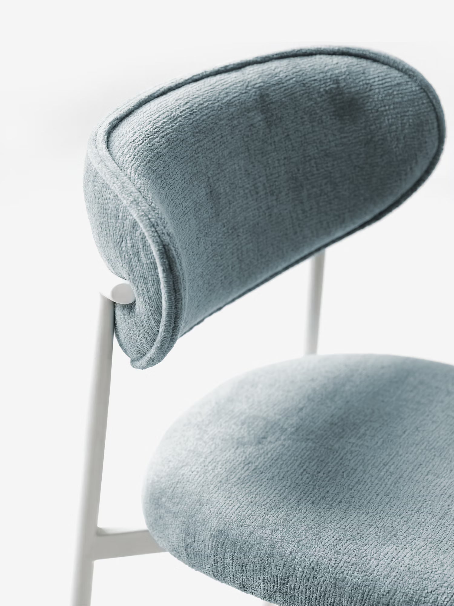Un primo piano della Luce Sedia imbottita di Divani.Store mette in risalto il suo morbido tessuto azzurro dalla trama ricca e lo schienale ricurvo. L'elegante struttura in metallo bianco crea un contrasto con il rivestimento, esaltando il suo design moderno e pulito su uno sfondo minimalista.