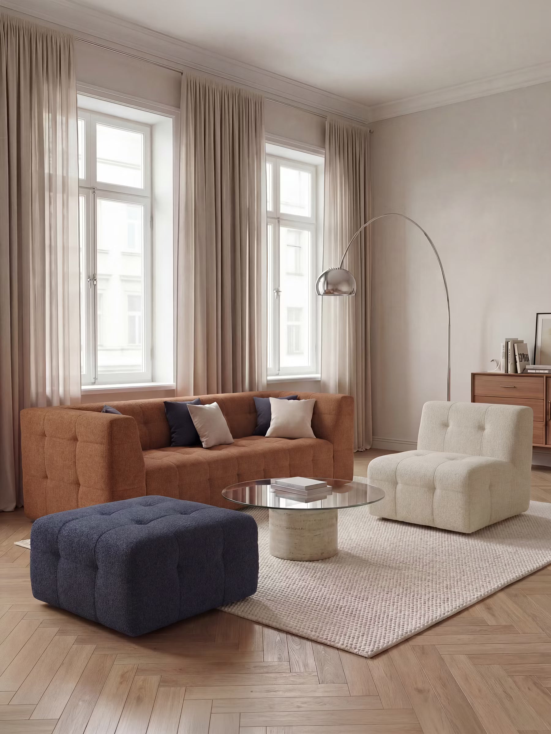 Soggiorno moderno con ampie finestre, tende beige, un divano componibile color ruggine, una poltrona capitonné in bouclé color crema e il pouf e modulo Mosaic in bouclé di Divani.Store — che misura 96x77 cm con rivestimento capitonné in tessuto T21 antimacchia — in blu navy. Lo spazio è caratterizzato da un tavolino rotondo in vetro, un tappeto chiaro, pavimento in legno, una lampada argentata ad arco e una credenza su cui sono esposti libri e oggetti decorativi, il tutto contro una parete chiara.