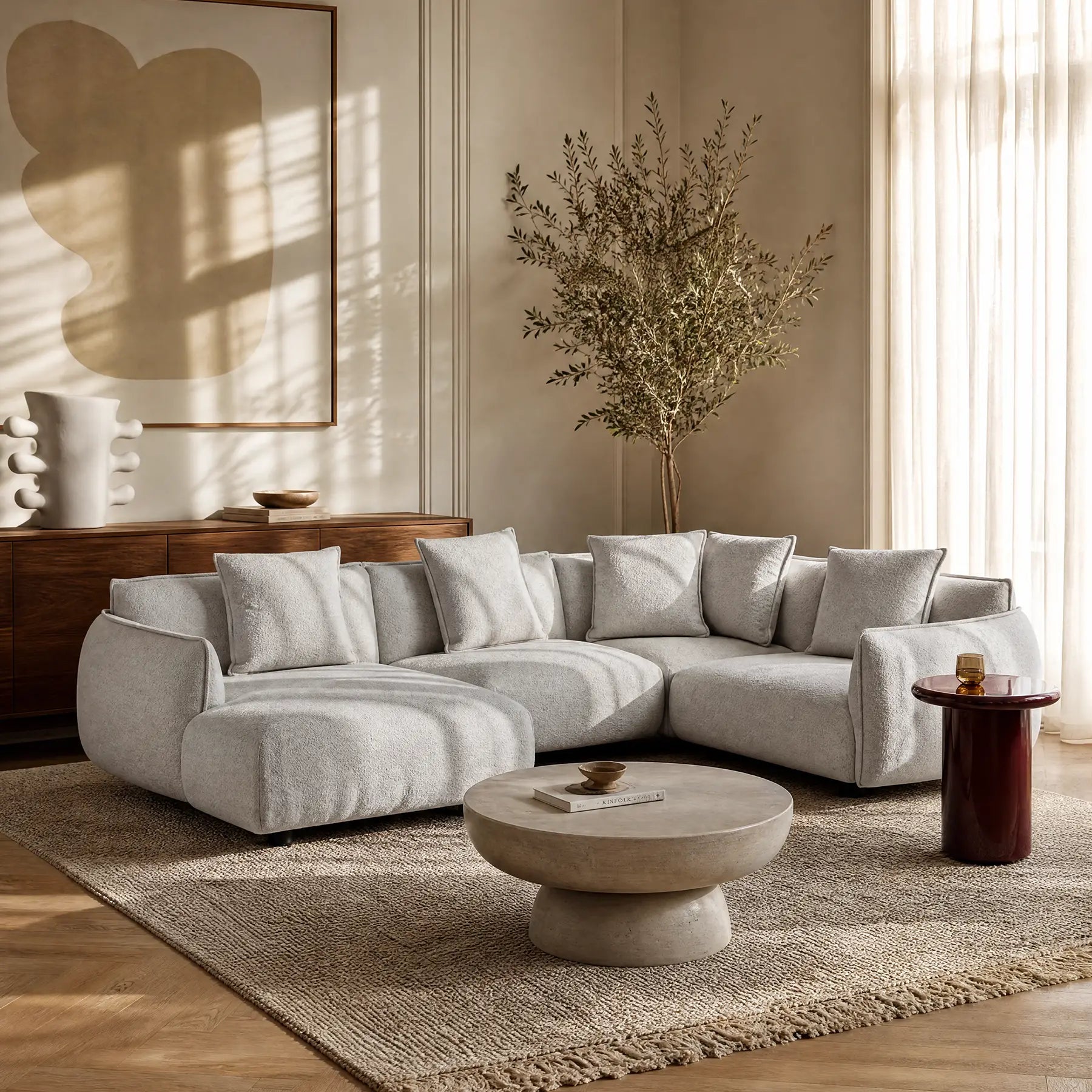 Soggiorno moderno con divano angolare panoramico componibile Nimbus con penisola e angolo in tessuto smacchiabile t33 di Divani.Store, su tappeto beige testurizzato, tavolino rotondo in pietra, credenza in legno, vasi in ceramica, arte astratta, pianta e tende velate.