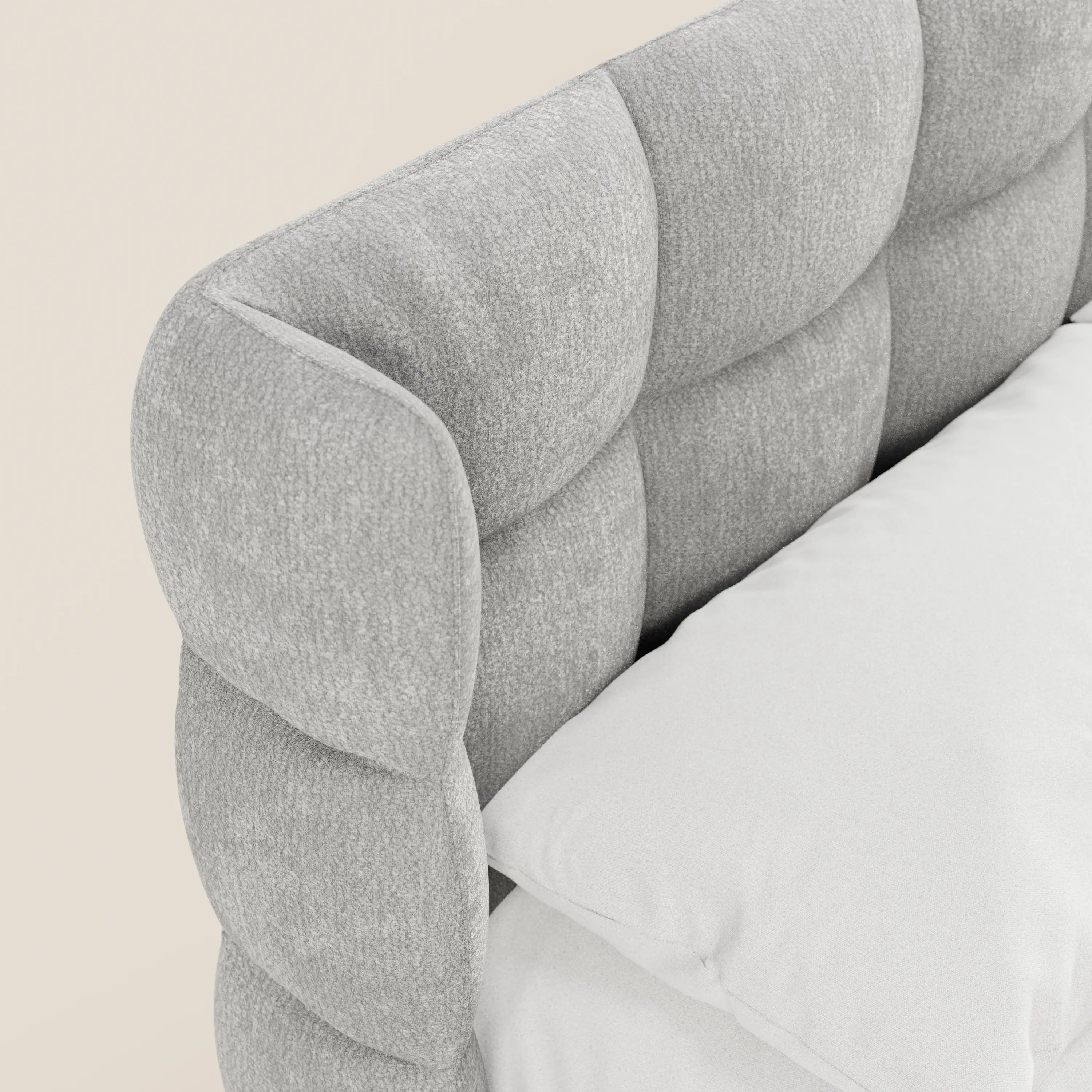 Primo piano del letto imbottito RAGNAR con contenitore e testiera capitonné avvolgente in elegante tessuto Oeko-Tex® di Letti.store. Un cuscino bianco e un coprimaterasso si stagliano contro la morbida testiera grigio chiaro, su uno sfondo beige neutro.