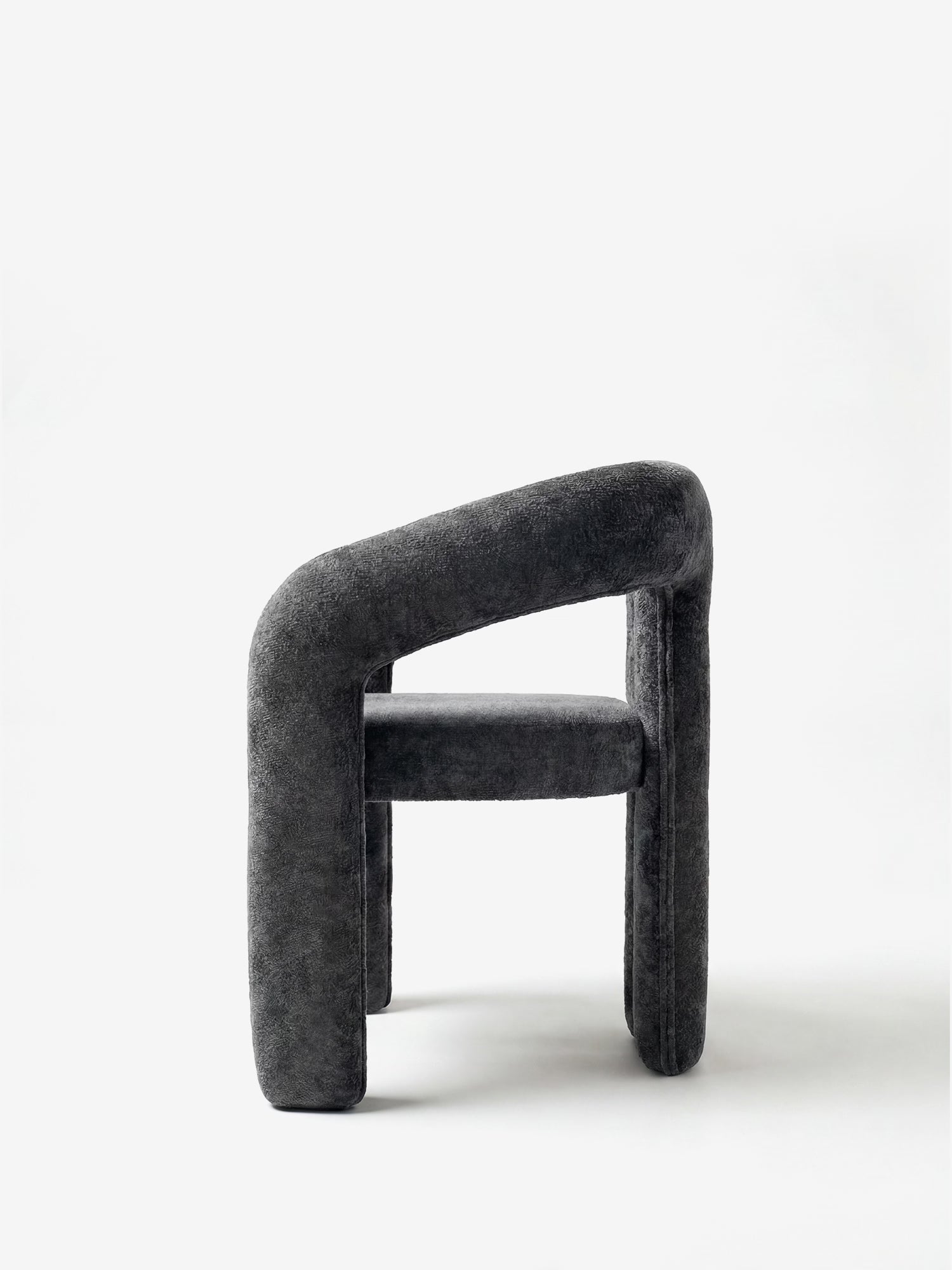 La sedia di design Sibilla di Divani.Store è una sedia moderna senza braccioli, di colore grigio scuro, interamente rivestita in tessuto, dalla forma sinuosa e con gambe e schienale spessi e arrotondati. Nella foto è raffigurata di profilo su uno sfondo bianco a tinta unita.