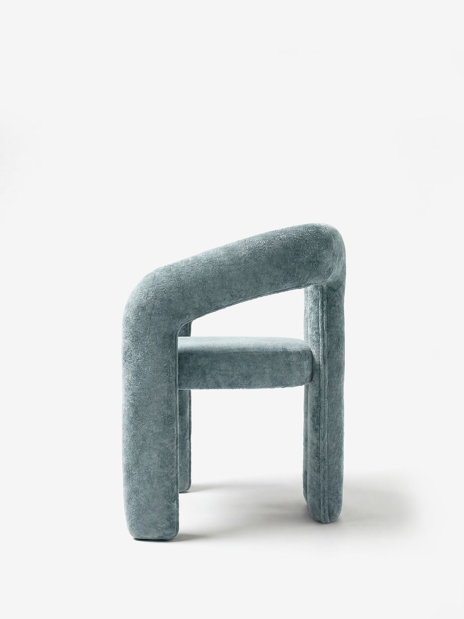 La sedia di design Sibilla di Divani.Store presenta una struttura ad arco interamente rivestita in morbido tessuto azzurro, gambe spesse e arrotondate e uno schienale curvo. Fotografata di profilo su sfondo bianco, mette in risalto il suo stile minimalista e scultoreo.