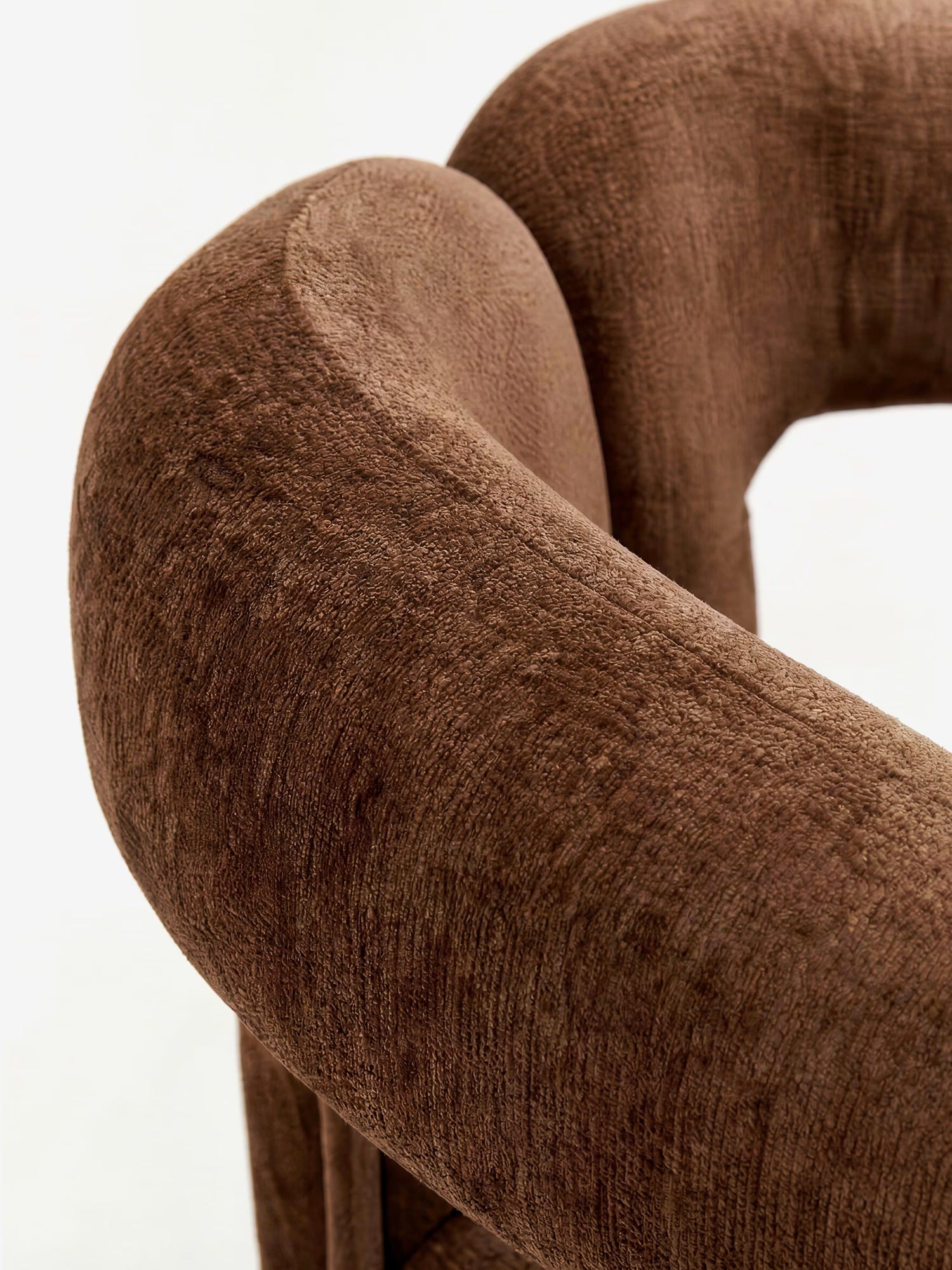 Primo piano della sedia di design Sibilla di Divani.Store: lo schienale curvo, interamente rivestito in morbido tessuto marrone, e la forma ad arco garantiscono una seduta accogliente e confortevole su uno sfondo neutro.