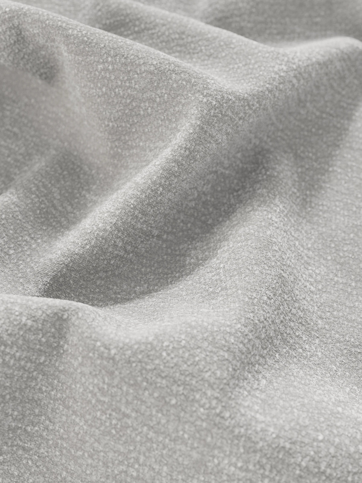 Un primo piano di un tessuto grigio chiaro dalla superficie tessuta e ruvida, drappeggiato in morbide pieghe su un letto imbottito ONYX con vano contenitore e testiera verticale in tessuto certificato Oeko-Tex®, con luci che ne esaltano la trama tattile.