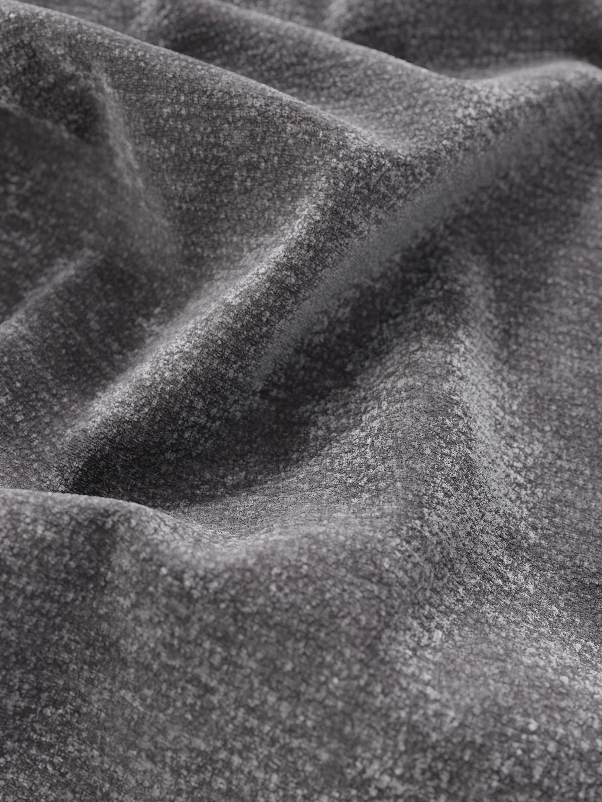 Un primo piano del tessuto grigio scuro del letto imbottito ONYX con contenitore e testiera verticale in tessuto certificato Oeko-Tex®, che mette in risalto la morbida trama, le delicate pieghe e i sottili giochi di luce e ombra sulla superficie intrecciata.