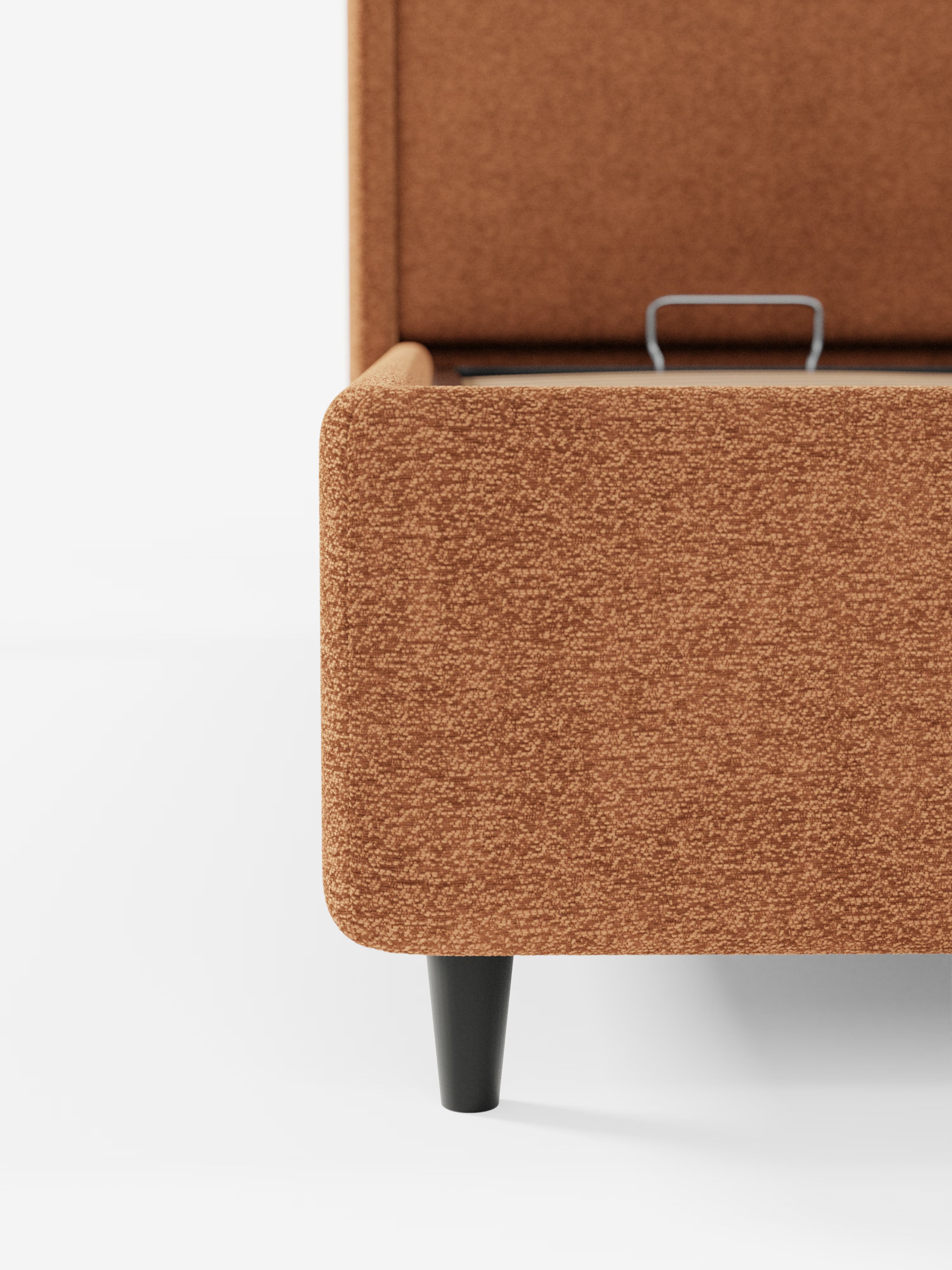 Primo piano del letto di design VALKIRIA con vano contenitore e testiera avvolgente di Letti.store, rivestito in tessuto Oeko-Tex® color ruggine. L’immagine mette in risalto un angolo arrotondato e una gamba corta nera del letto contenitore, con il coperchio parzialmente aperto che lascia intravedere una maniglia in metallo all’interno. Sono presenti anche due cuscini imbottiti. Lo sfondo è bianco a tinta unita.