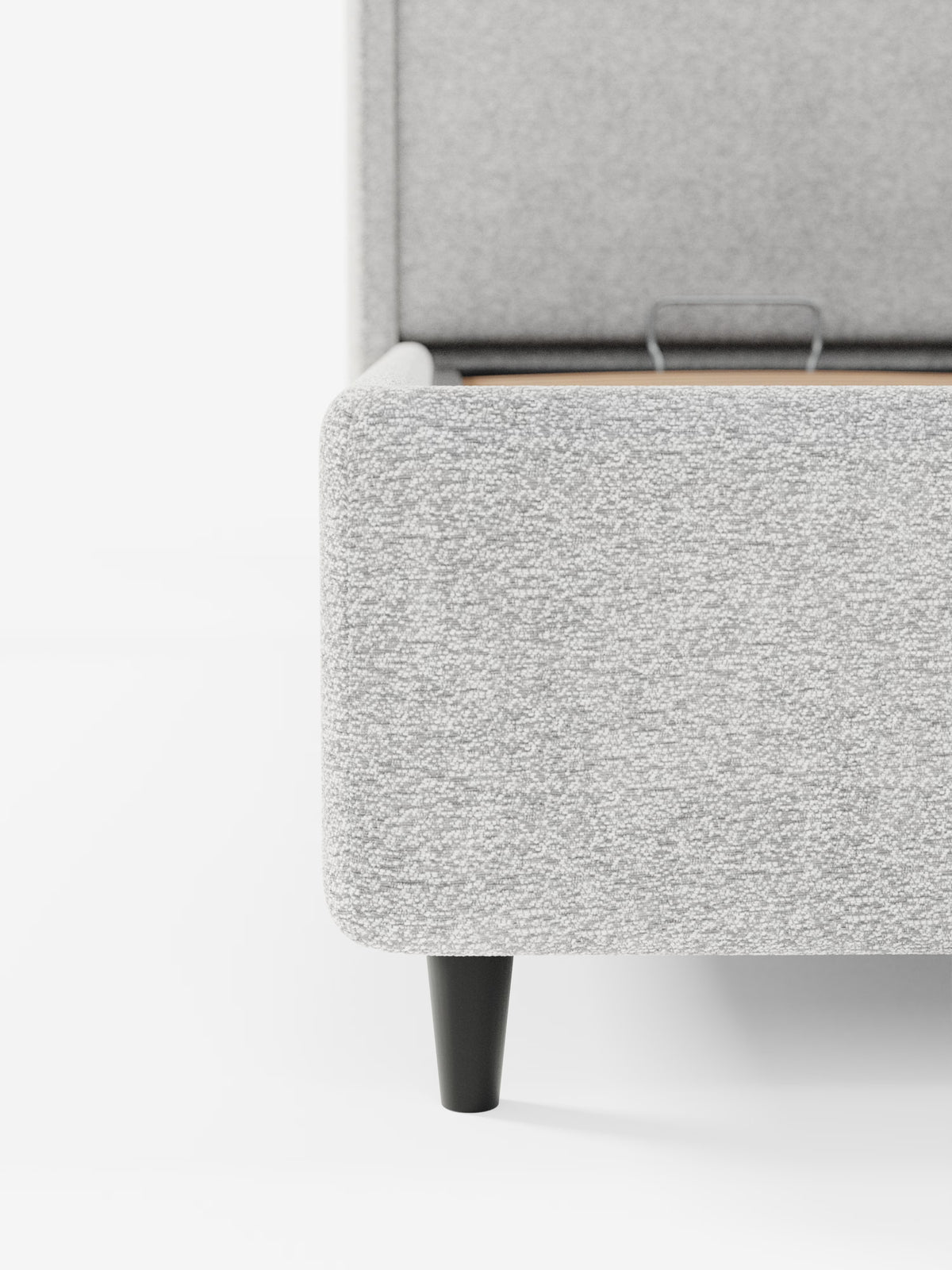 Primo piano dell'angolo di un letto di design VALKIRIA con vano contenitore e testiera avvolgente di Letti.store, rivestito in morbido tessuto Oeko-Tex® dalla trama ruvida, color grigio chiaro. La struttura contemporanea è caratterizzata da due cuscini imbottiti e poggia su eleganti gambe affusolate di colore nero. Le doghe del letto e una staffa di sostegno in metallo sono parzialmente visibili su uno sfondo bianco a tinta unita.