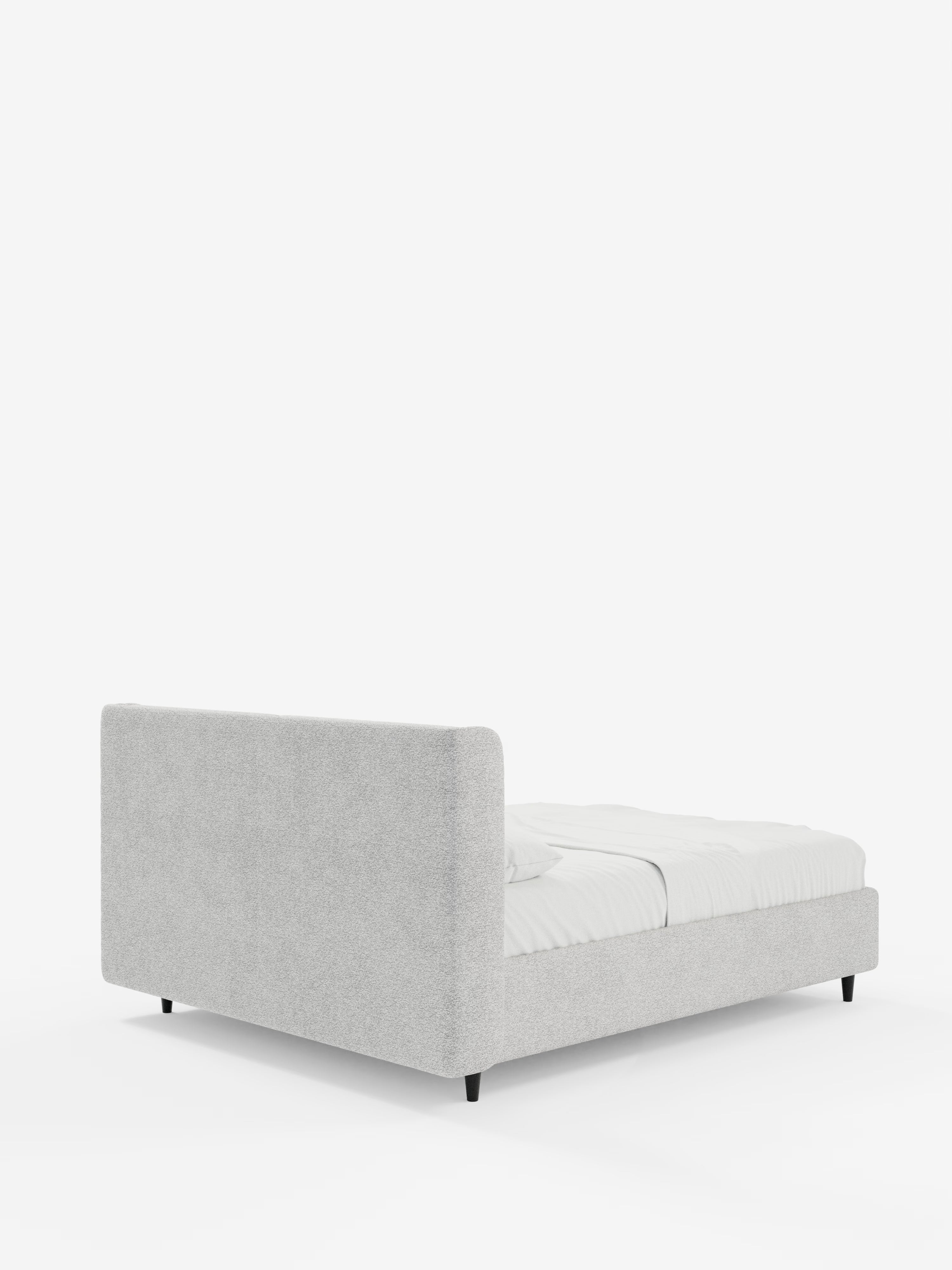 Il letto di design VALKIRIA con contenitore e testiera avvolgente di Letti.store presenta una testiera alta e imbottita di colore grigio chiaro, con due morbidi cuscini in tessuto Oeko-Tex® e fianchi bassi, il tutto sostenuto da corte gambe nere. Il materasso bianco è parzialmente coperto da un lenzuolo bianco drappeggiato in modo morbido, mentre lo sfondo bianco a tinta unita mette in risalto il design contemporaneo e la texture raffinata del letto.